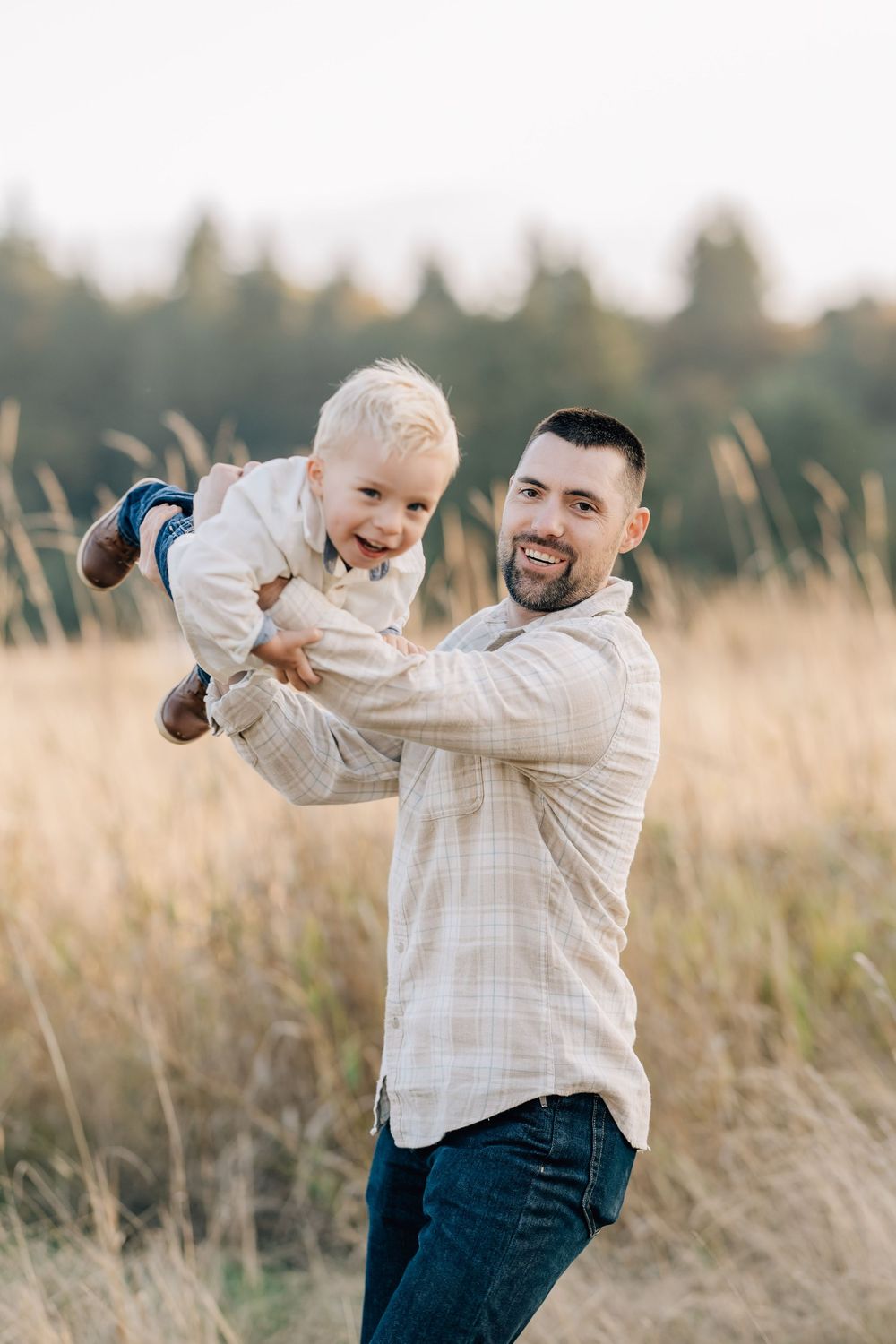 A smiling father lifts his happy toddler son in a golden field with trees in the background.