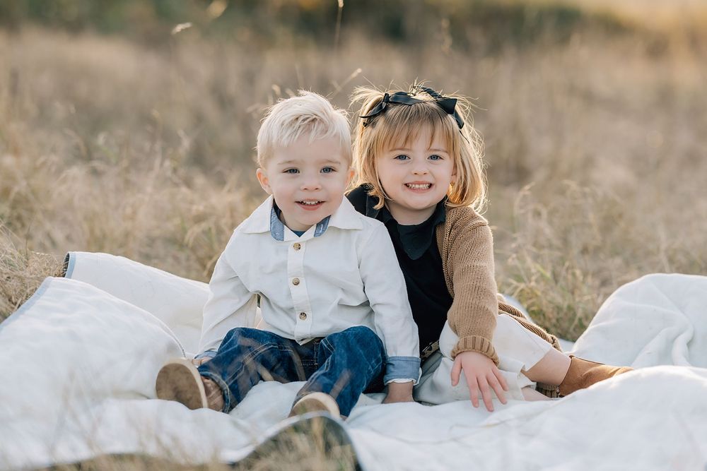 Two young children smiling and sitting together on a white blanket in a golden field outdoors.