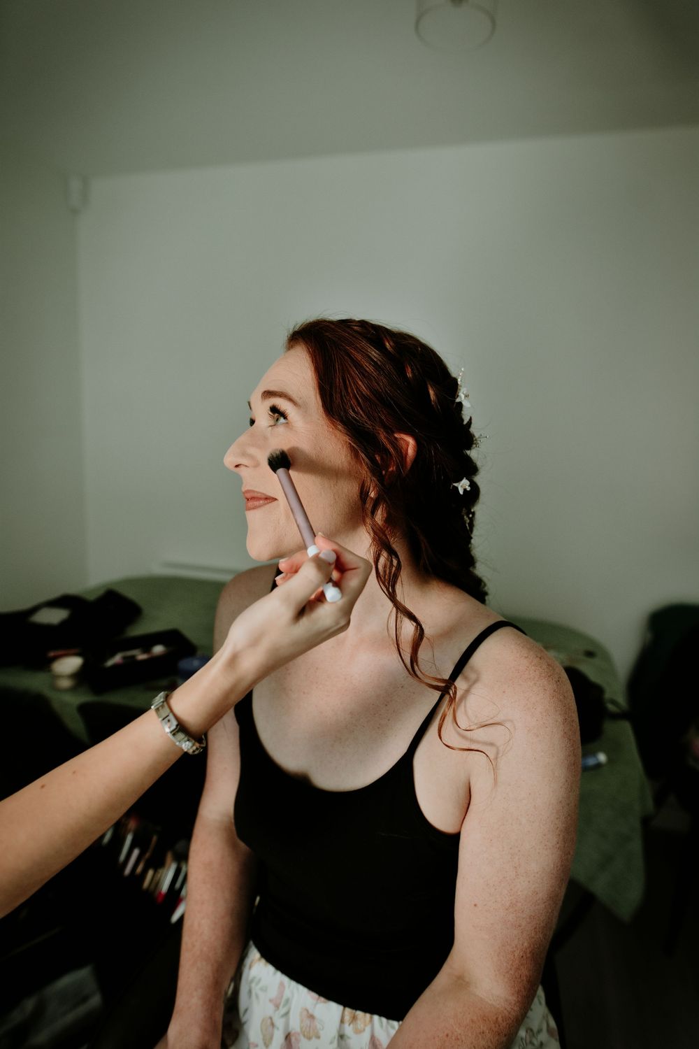 Bride with red hair smiling as makeup artist applies blush with brush during wedding morning preparations.