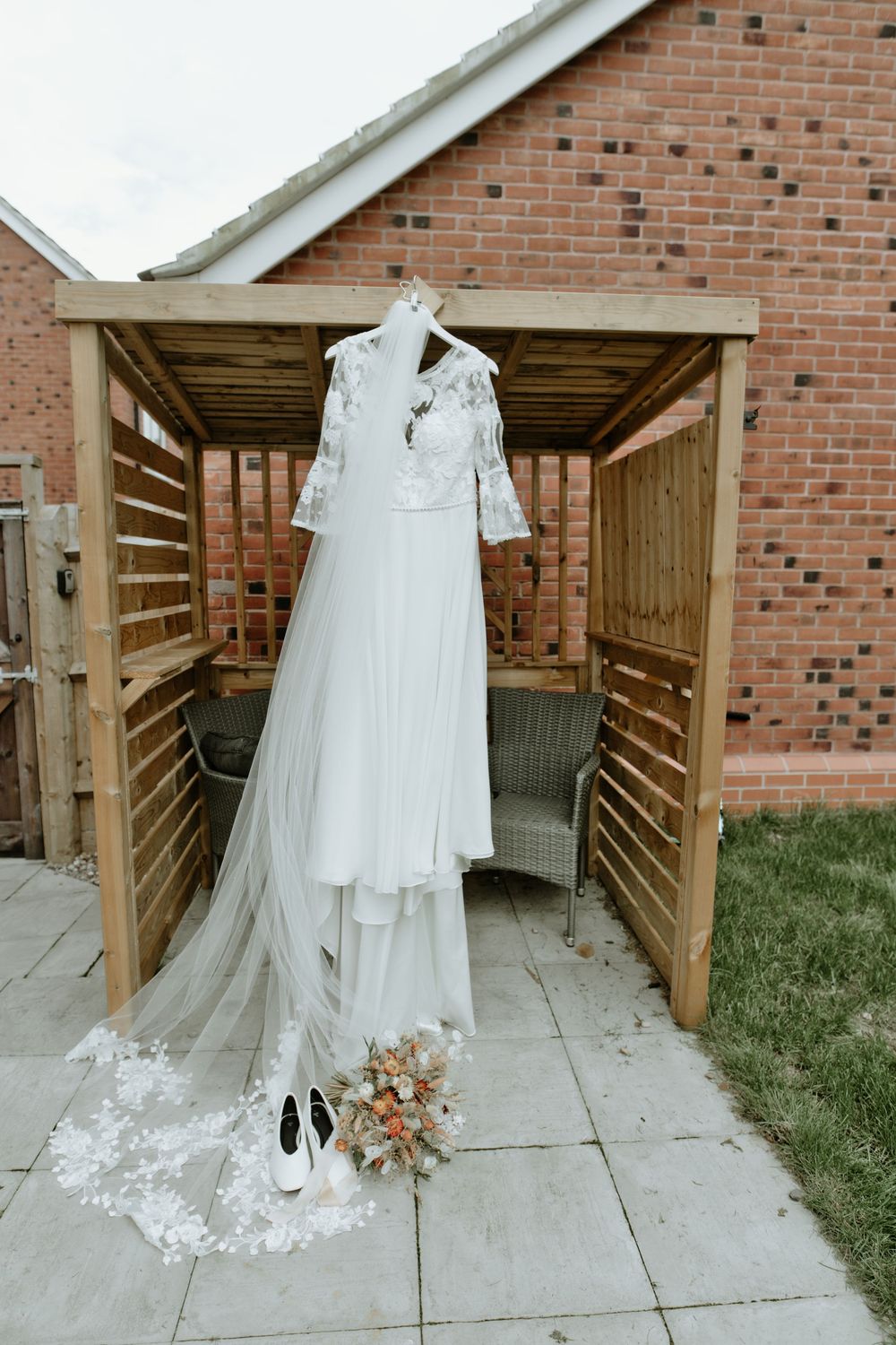White wedding dress with lace bodice and veil displayed on mannequin outdoors with bridal bouquet and shoes below.