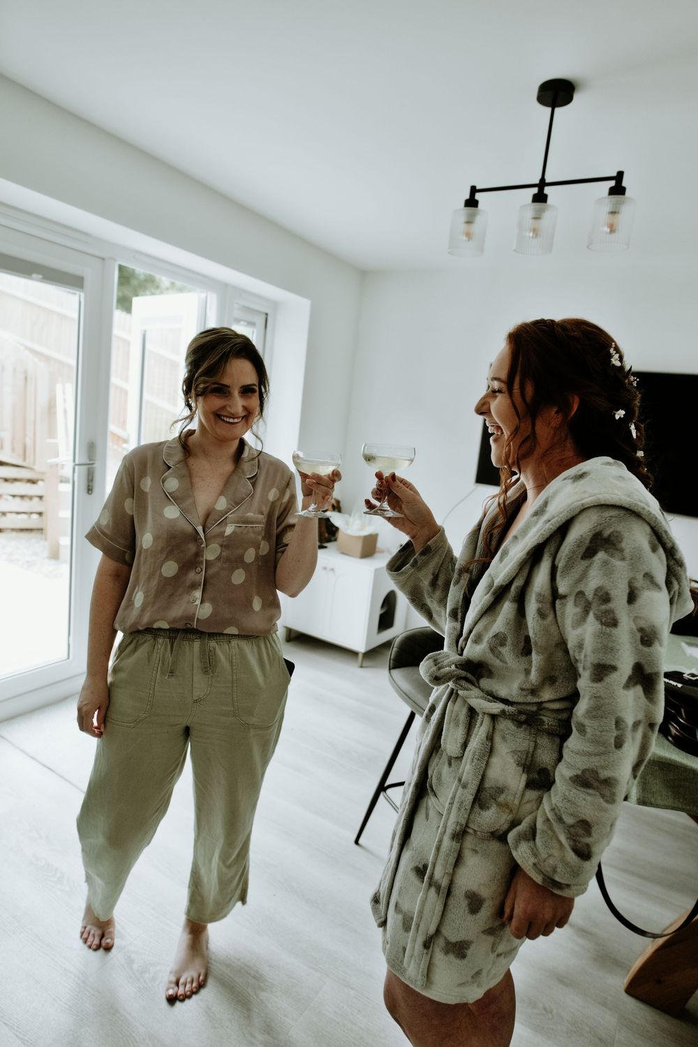 Two bridesmaids in robes toasting with champagne glasses while getting ready for a wedding.
