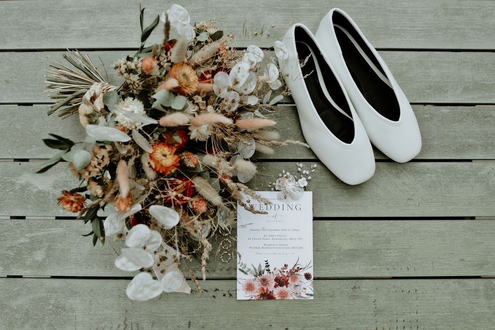 Boho wedding flat lay featuring a dried floral bouquet, white ballet flats, and an elegant floral wedding invitation on wood.