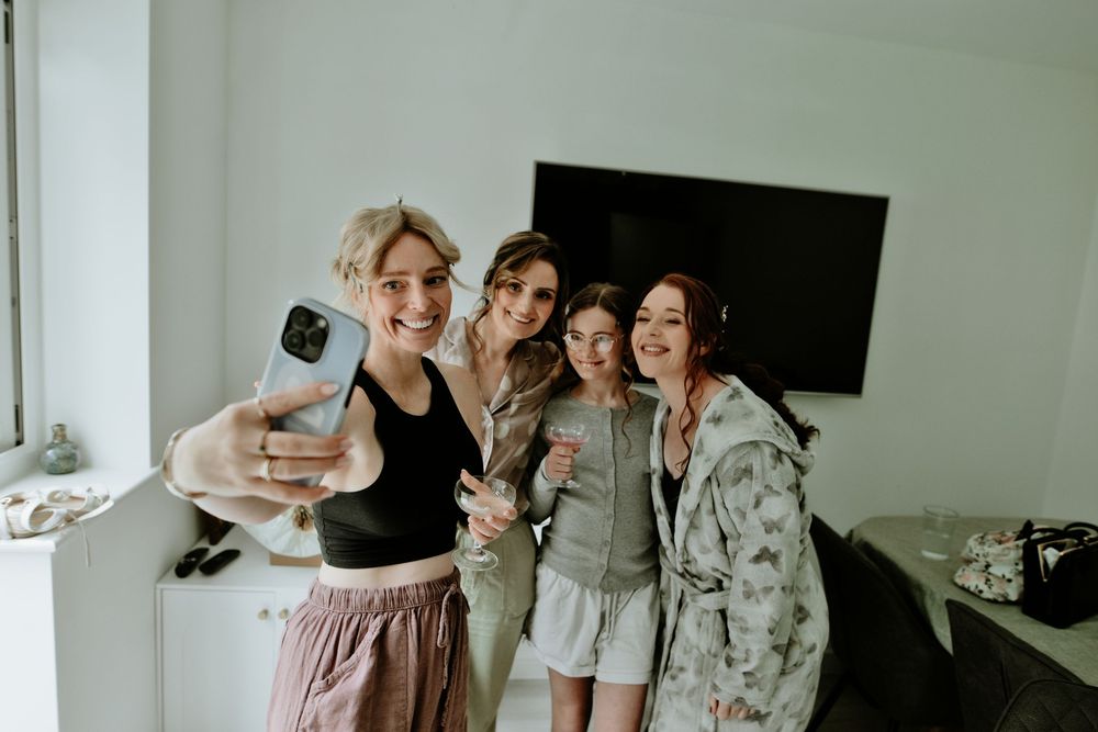 Four women smile together taking a selfie indoors, dressed casually in a bright modern room.