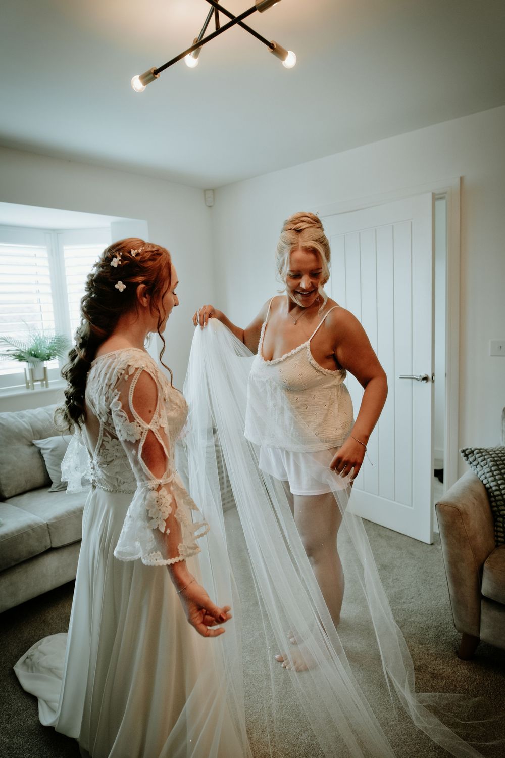 Bride in lace wedding dress getting veil fitted by bridesmaid in white lingerie in bright living room.