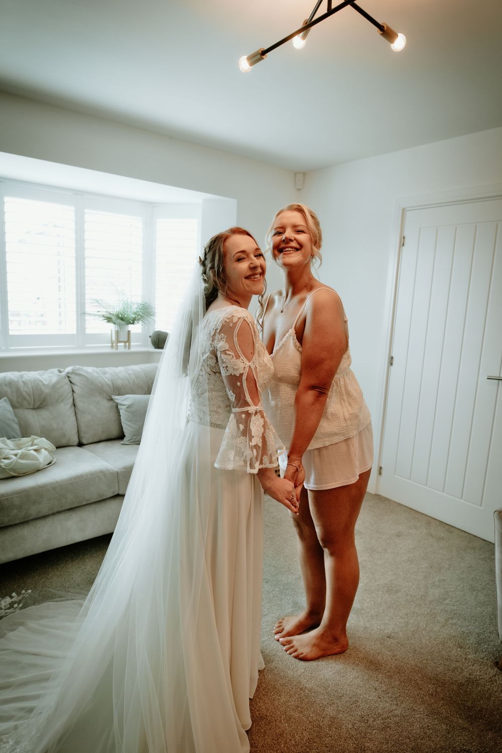 Bride in lace gown and veil poses smiling with bridesmaid in white lingerie in cozy living room.
