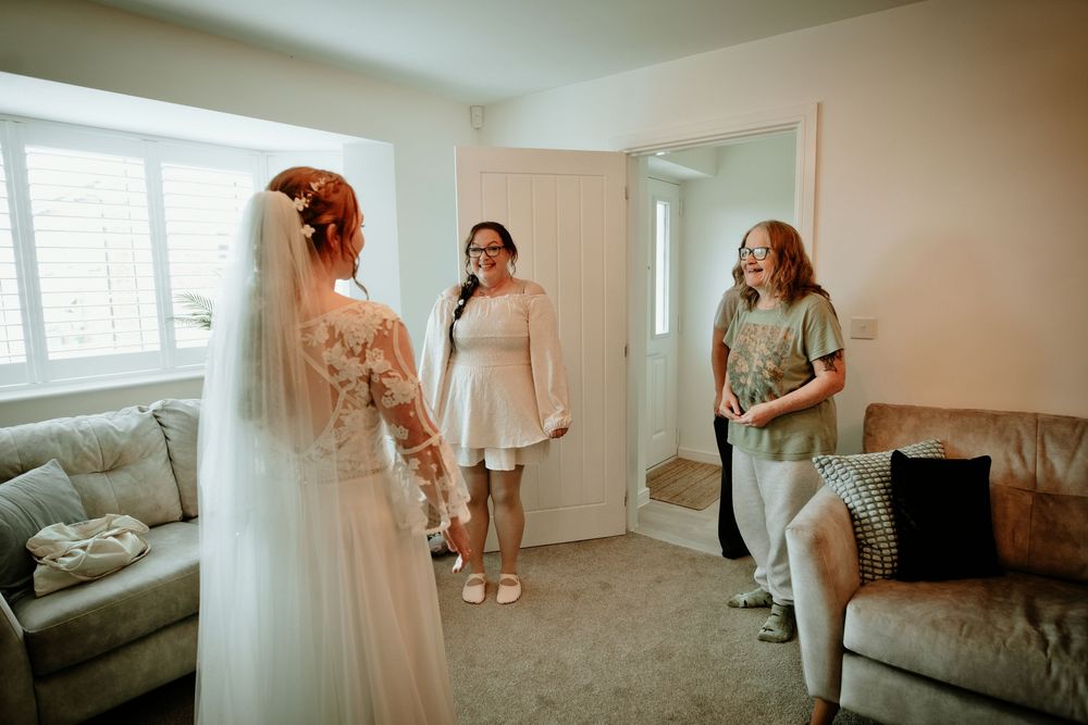 A bride in a white gown and veil reveals her look to emotional bridesmaids at home.