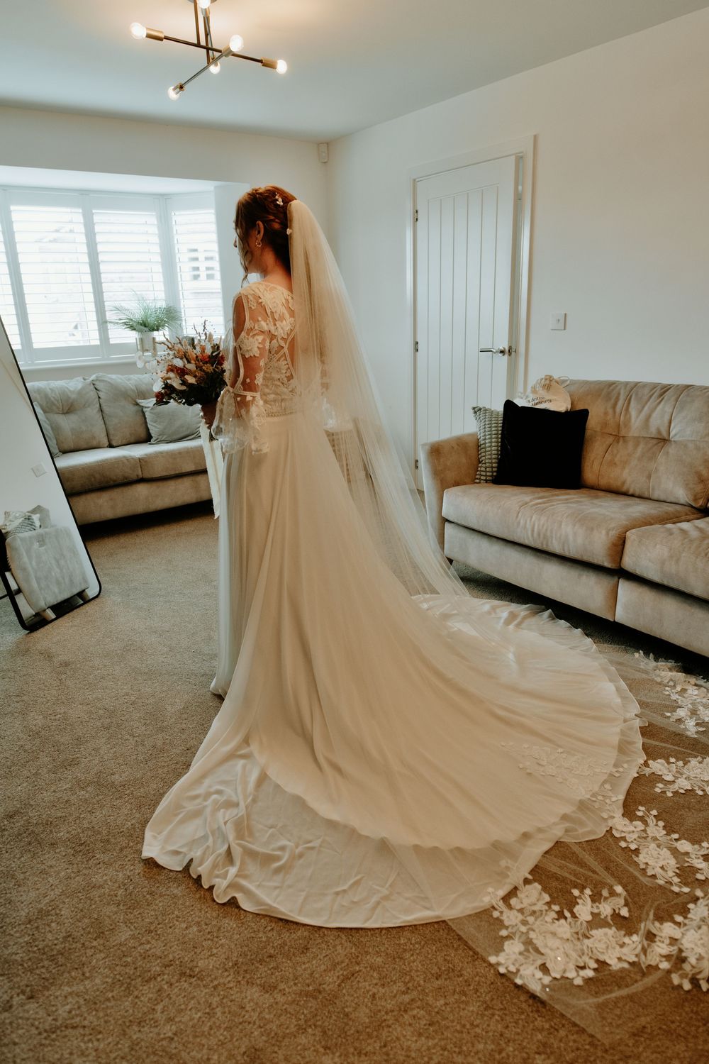 Bride in elegant lace wedding gown with long cathedral veil and floral trim stands in bright living room.