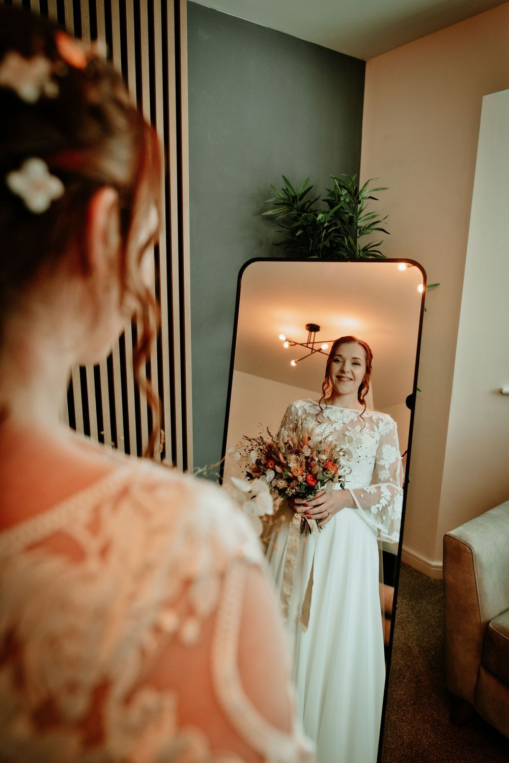 Radiant bride in lace gown holding colorful bouquet smiling at her reflection in a full-length mirror.