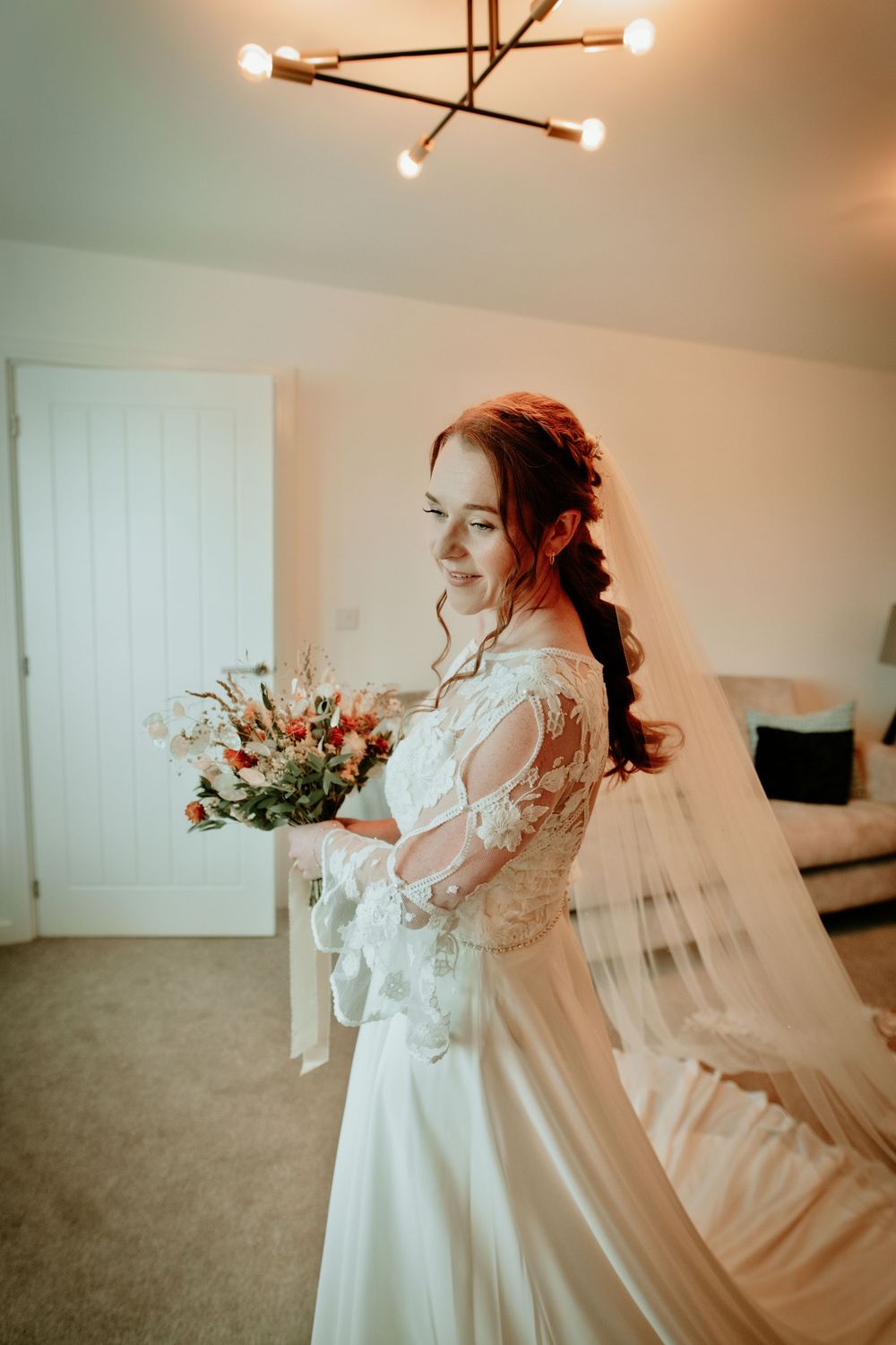 Bride in lace wedding dress holding autumn bouquet with veil flowing, smiling in a bright room.