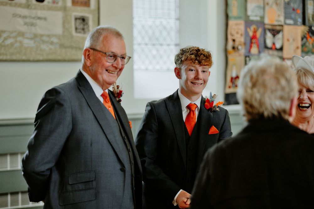 Elderly man and teenage boy in suits with orange boutonnieres smiling at wedding ceremony.