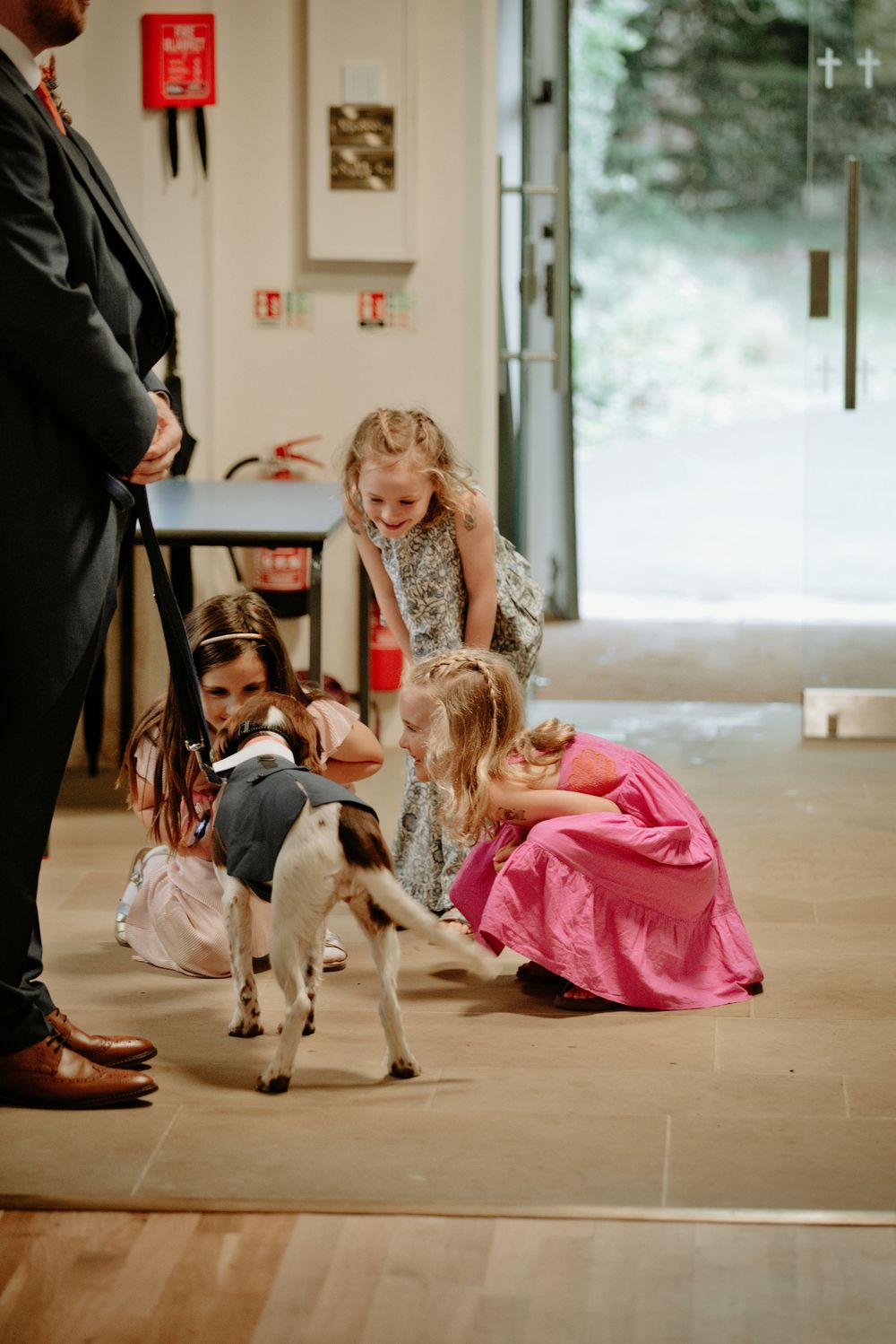 Children in formal attire pet a baby goat dressed in a suit at a wedding venue.