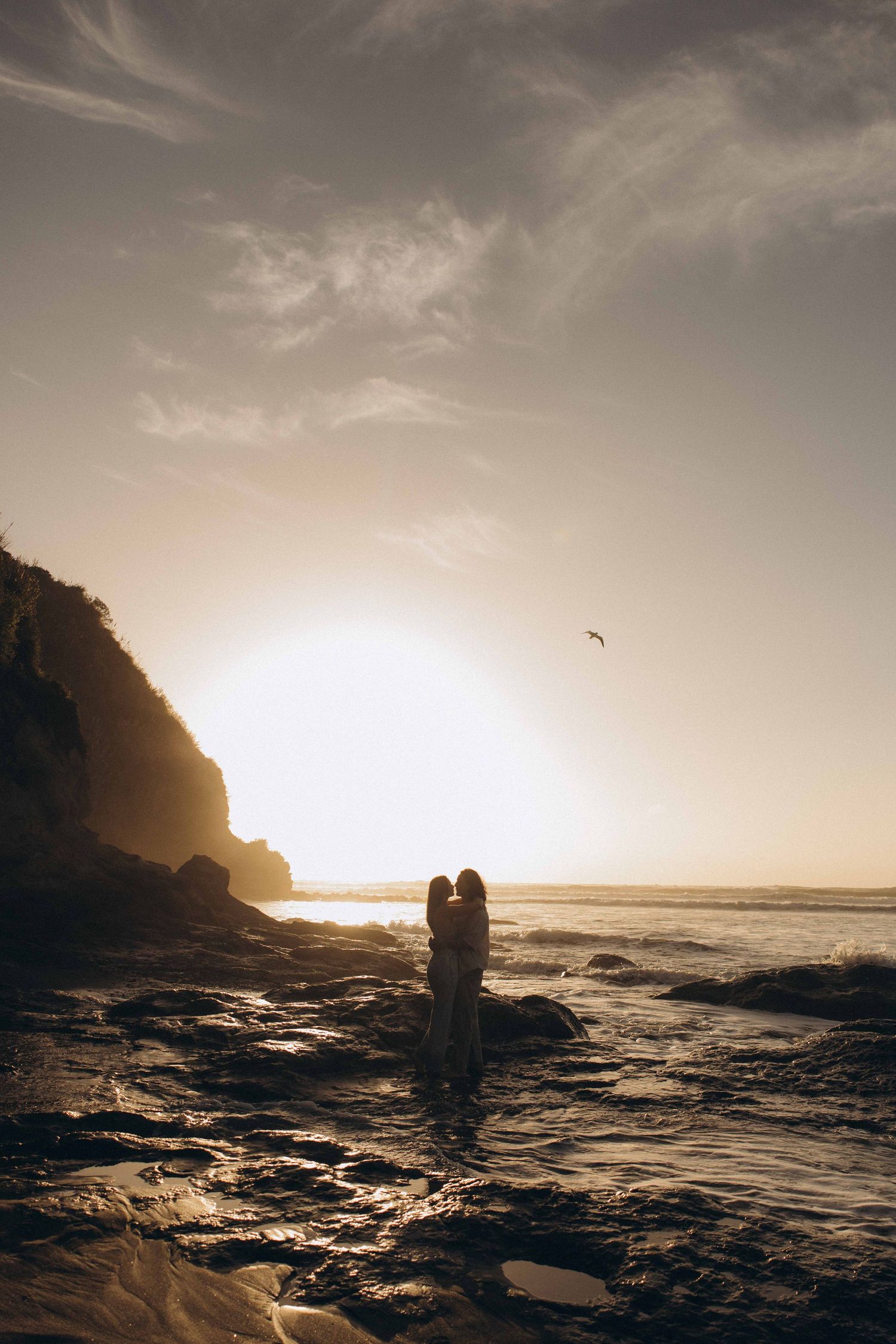 Silhouette of couple embracing on rocky beach at golden sunset with ocean waves and cliff backdrop.