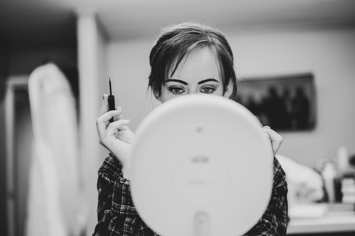 Black and white photo of a woman applying mascara while looking into a round mirror.