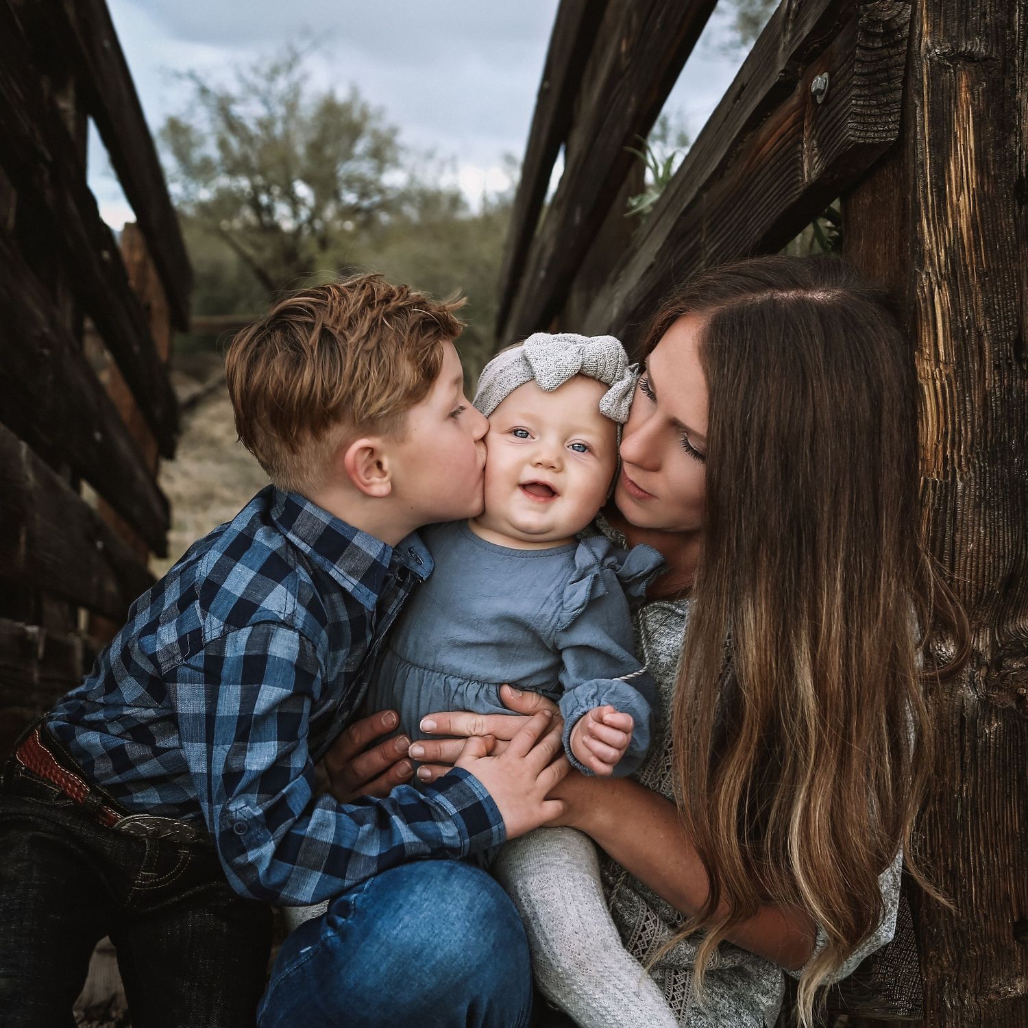 A boy kisses the cheek of a smiling baby in a gray bow headband as their long-haired mother holds them near rustic weathered wood.