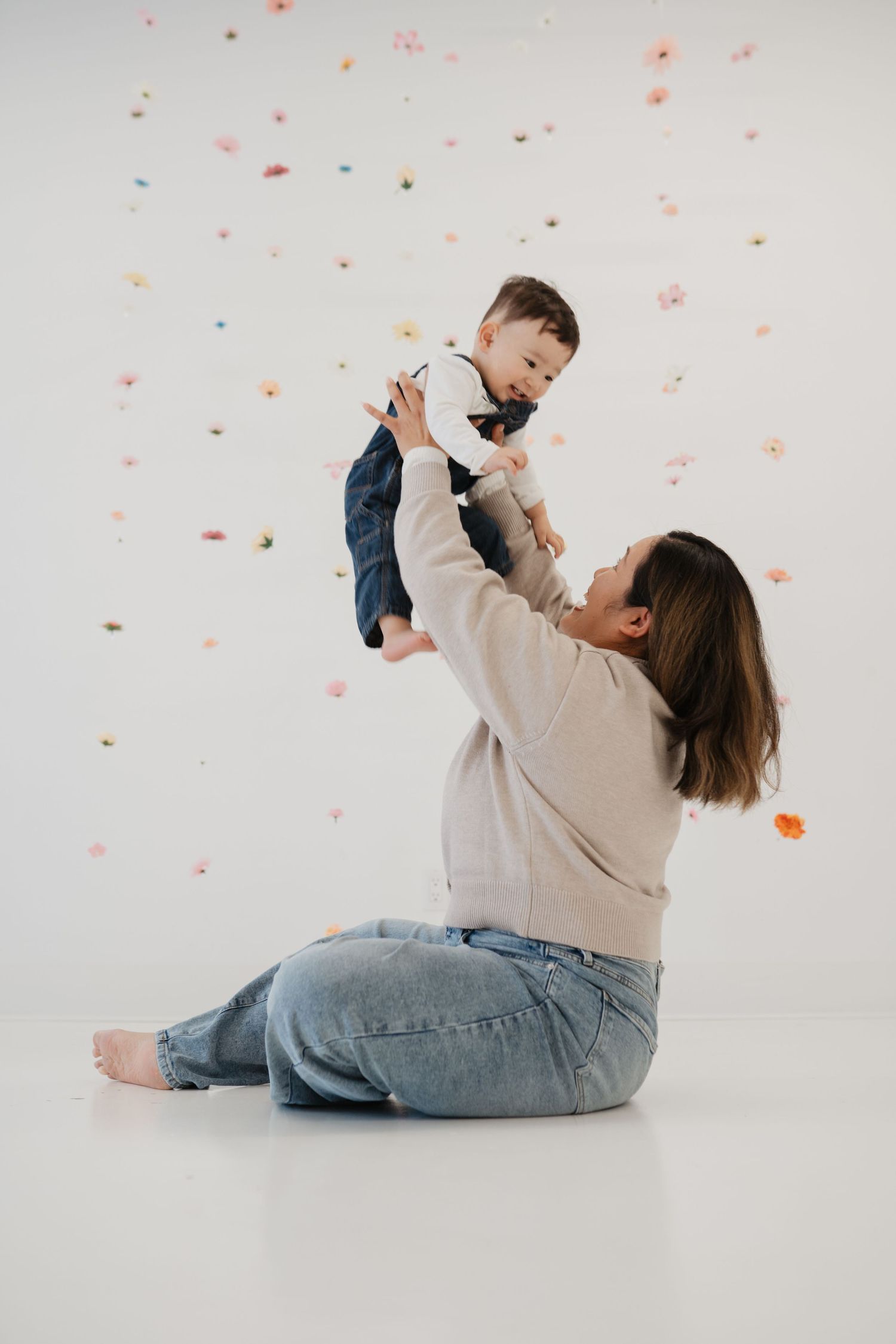 Baby in denim overalls crawling on white floor toward floral wall decor with scattered pink flowers.
