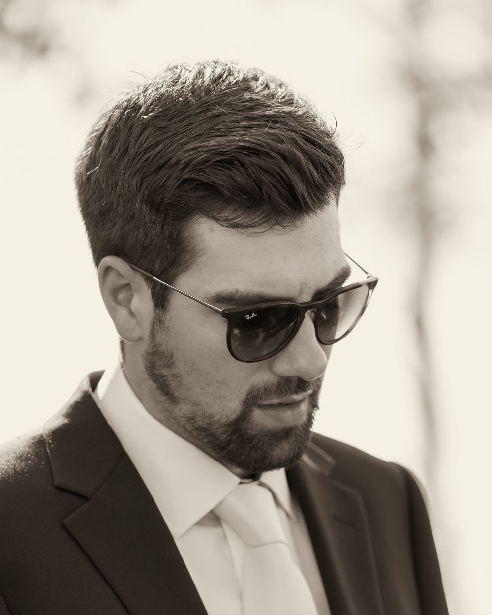Stylish groom in black suit wearing Ray-Ban sunglasses in black and white portrait photo.