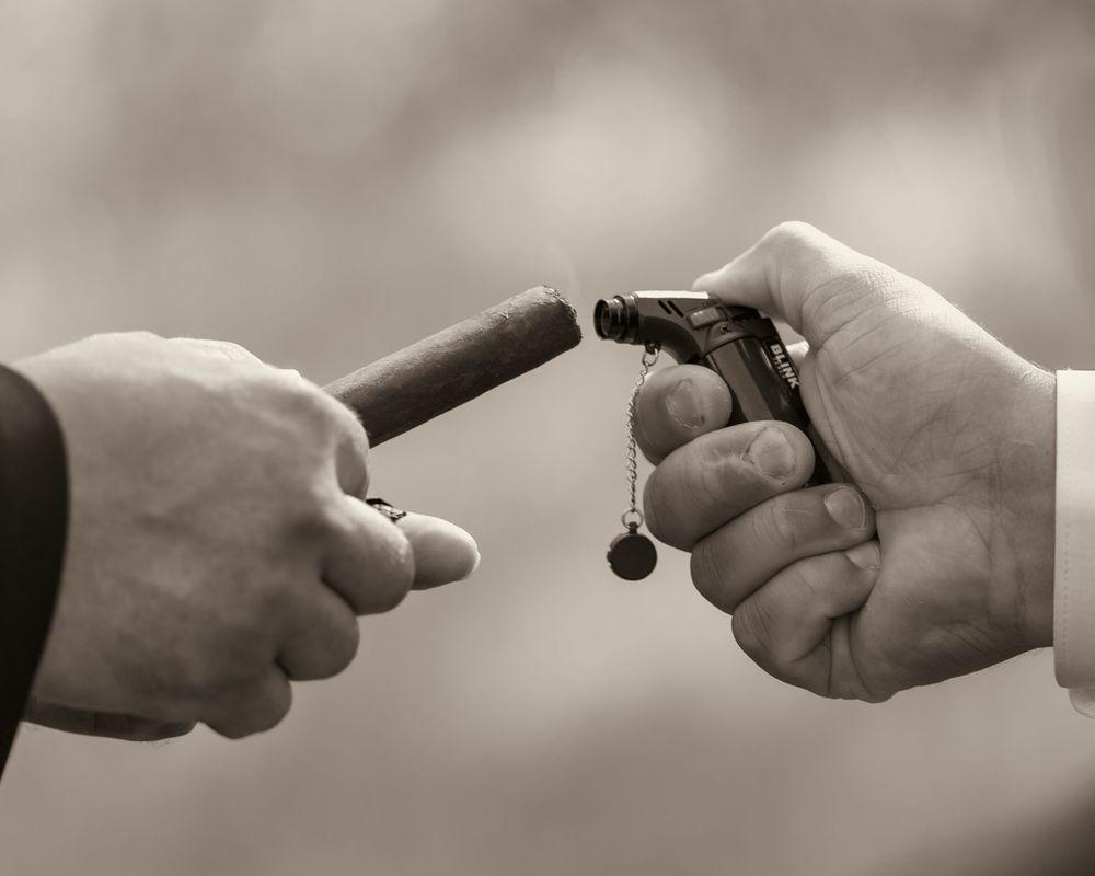 Close-up black and white photo of hands lighting a cigar with a torch lighter at wedding.