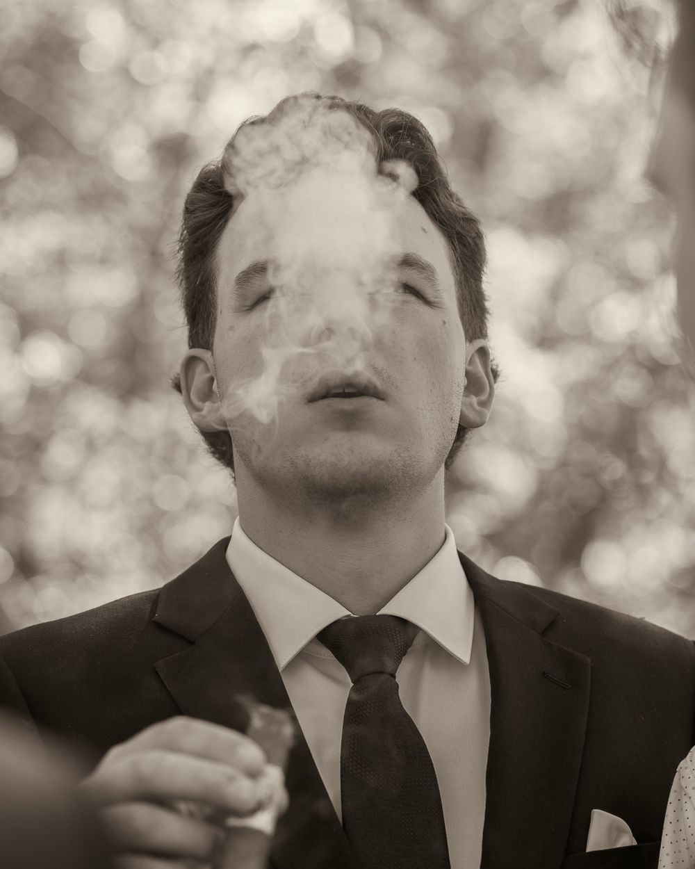 Groom in black suit exhaling cigar smoke in artistic black and white wedding portrait.