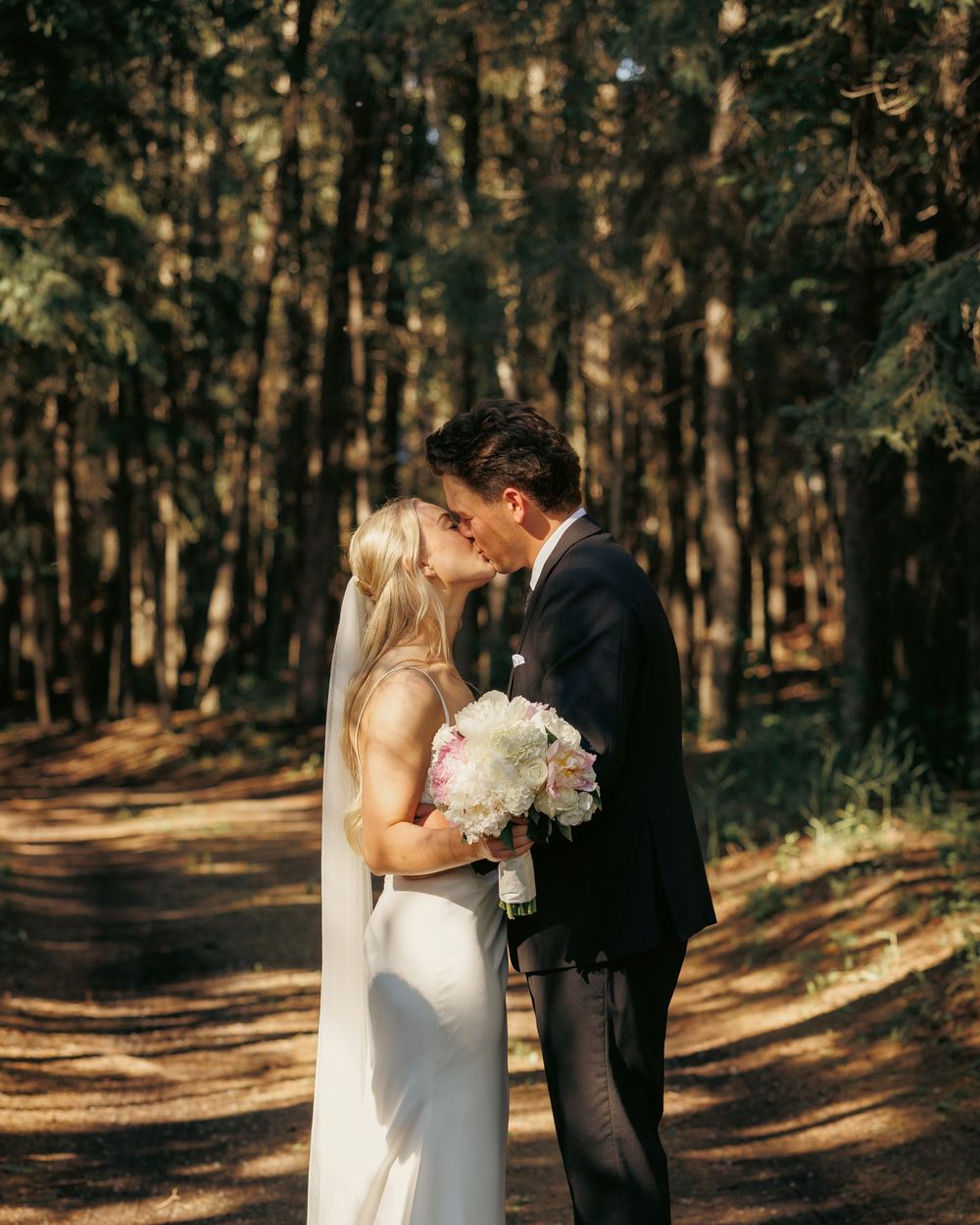 Bride and groom sharing first kiss on forest path, bride holding white bouquet in sunlight.