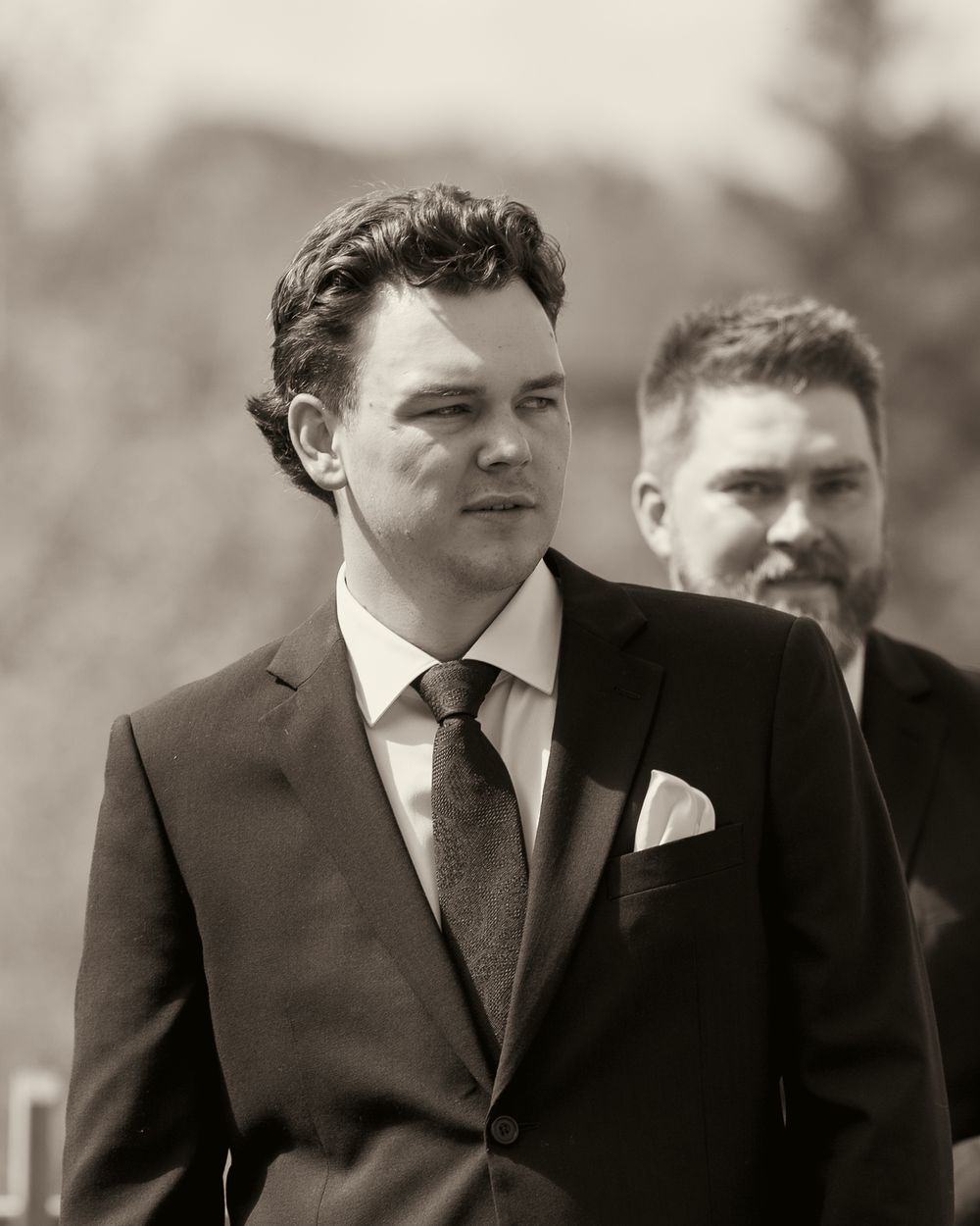 Black and white portrait of a dapper groom in a suit and tie at an outdoor wedding.