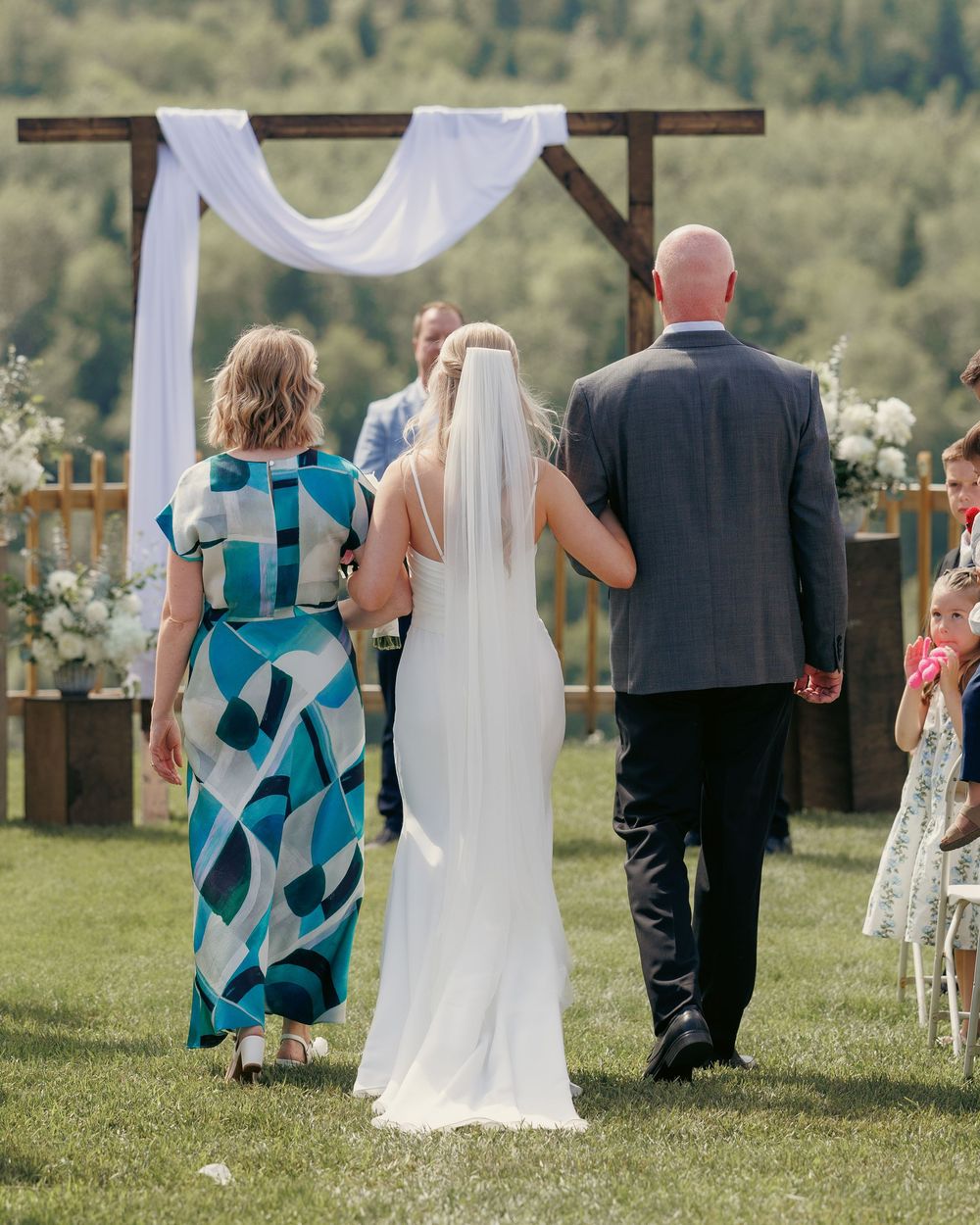 Bride walking down the aisle with parents toward a rustic wooden arch at outdoor wedding.