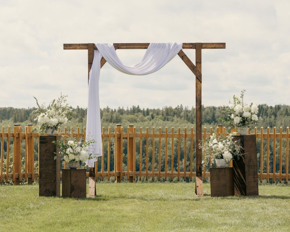 Rustic wooden wedding arch with white draping and floral arrangements on a green lawn.