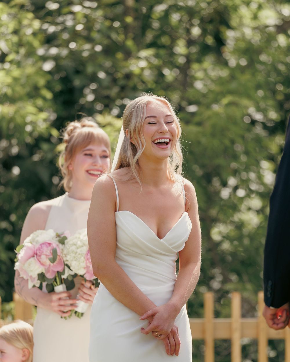 Joyful bride in a white spaghetti strap gown laughing during outdoor wedding ceremony.