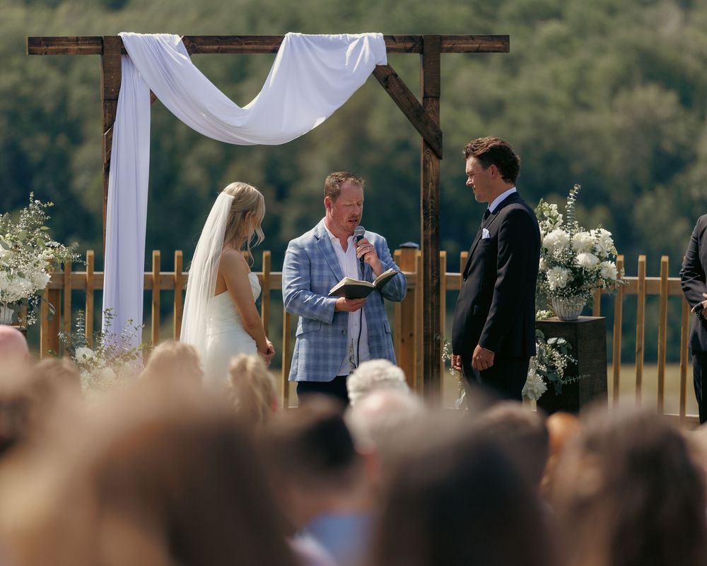 Outdoor wedding ceremony under rustic wooden arch with white draping, officiant reading vows to bride and groom.