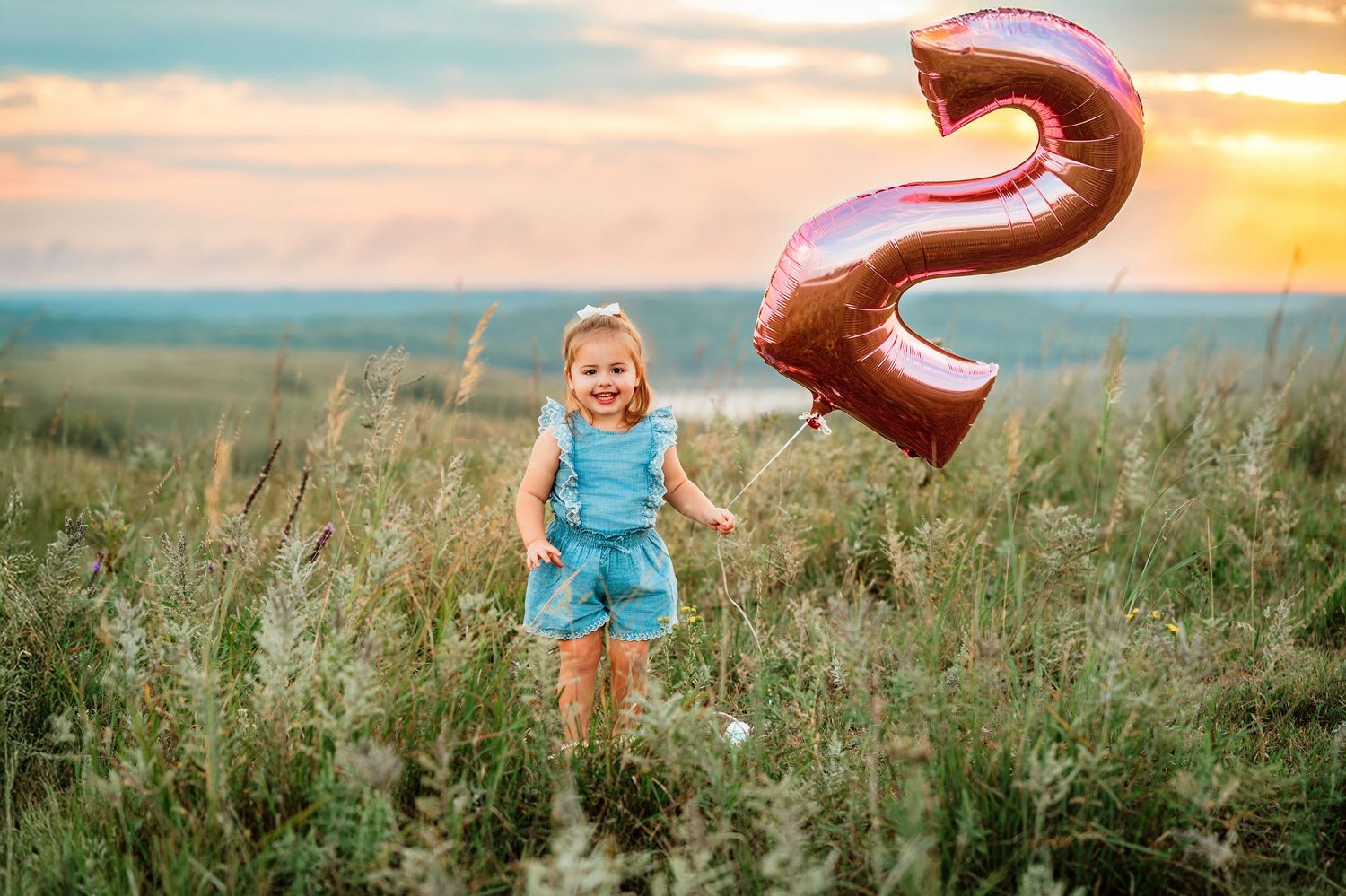 Smiling toddler girl holding a pink number 2 balloon in a field at sunset, celebrating her 2nd birthday.