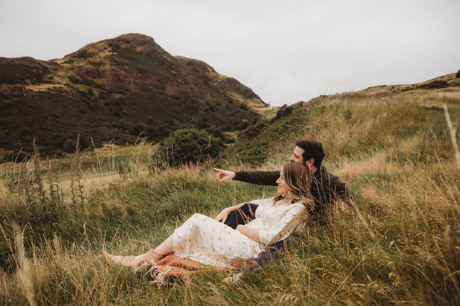 Couple sitting together in lush green hillside meadow, woman in white lace dress pointing toward scenic mountain landscape.