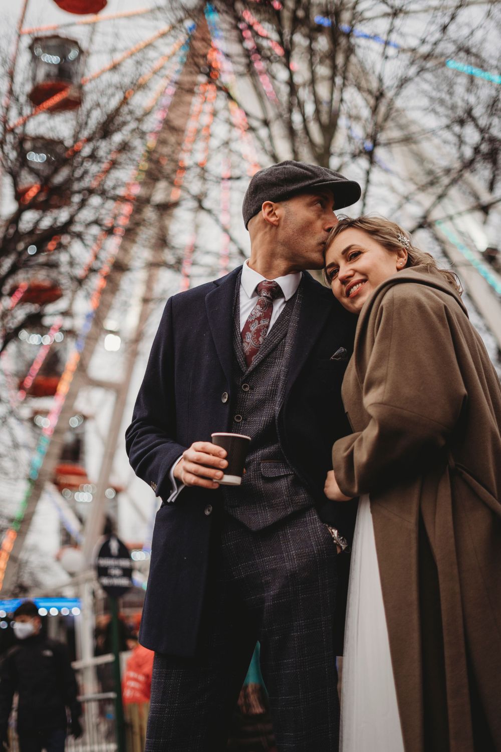 A couple in vintage attire shares a tender moment near a ferris wheel at twilight.