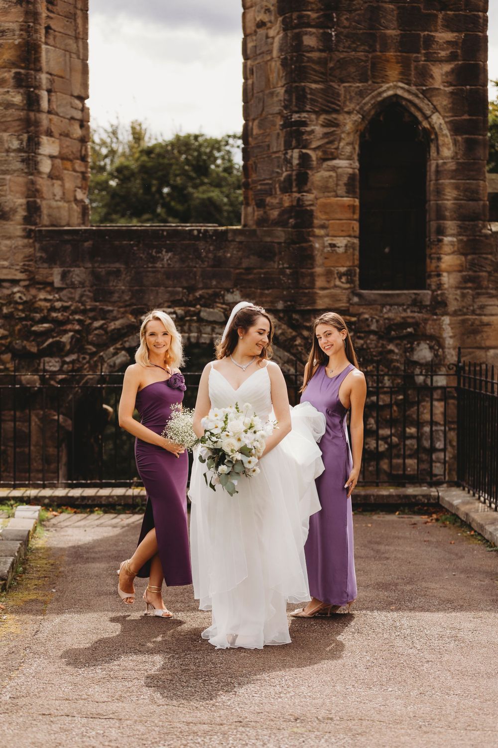 Bride and bridesmaids in purple dresses pose against stone church ruins.