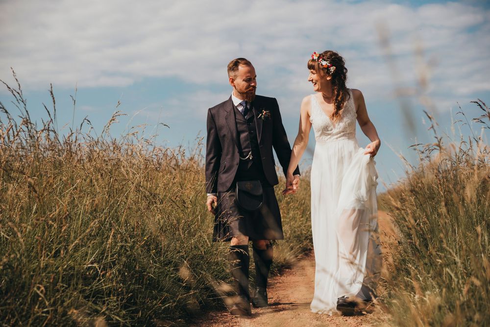 A couple walks hand in hand along a dirt path through tall grass on their wedding day.