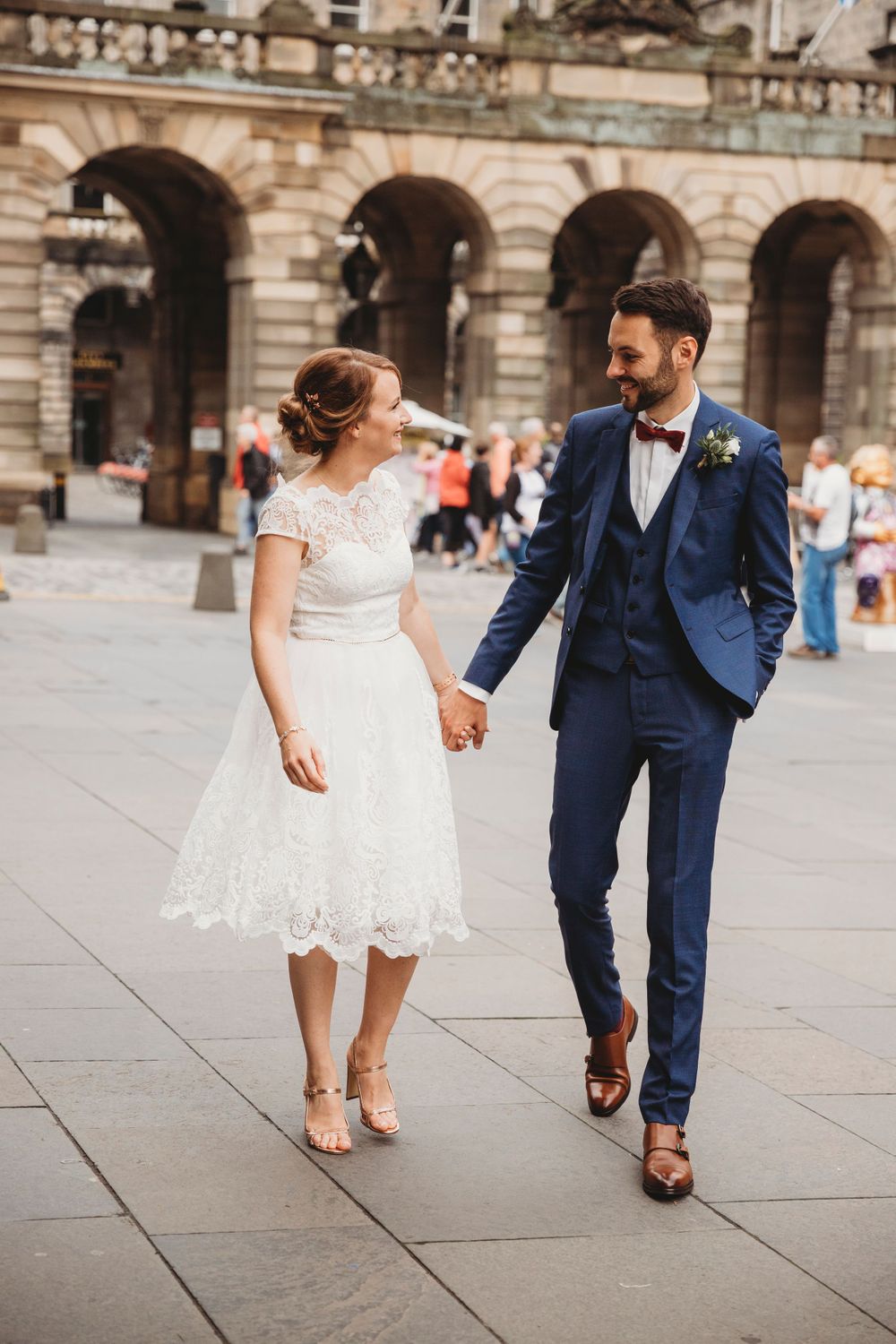 Wedding couple walks together on stone plaza with historic architecture in background.