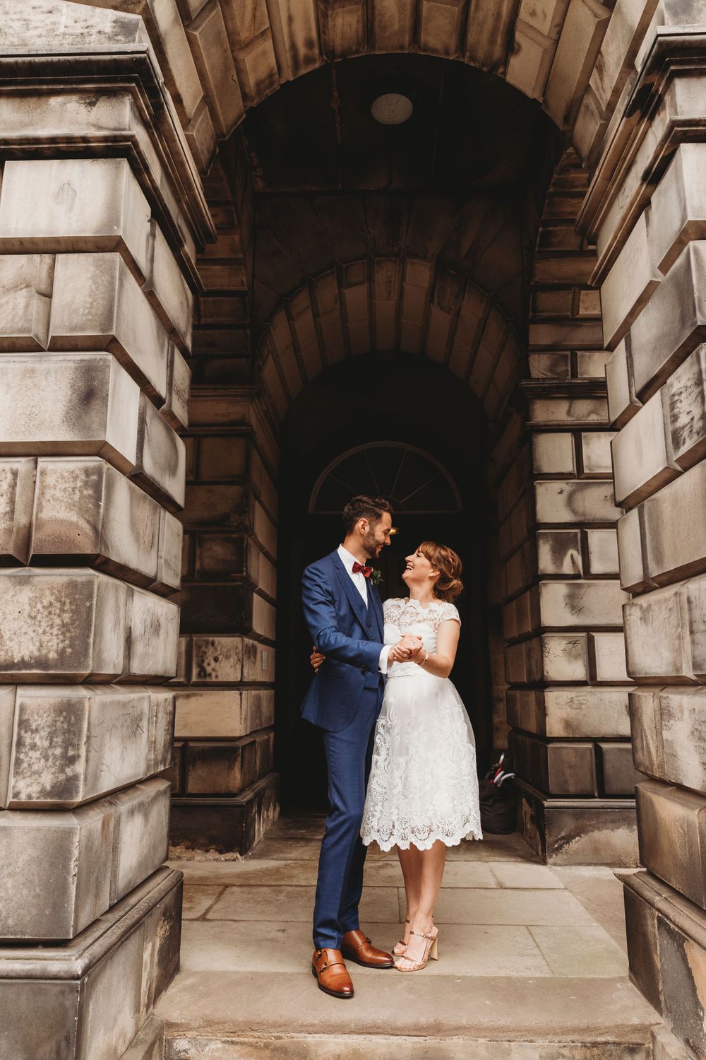 Couple in wedding attire stand together under arched stone passageway.