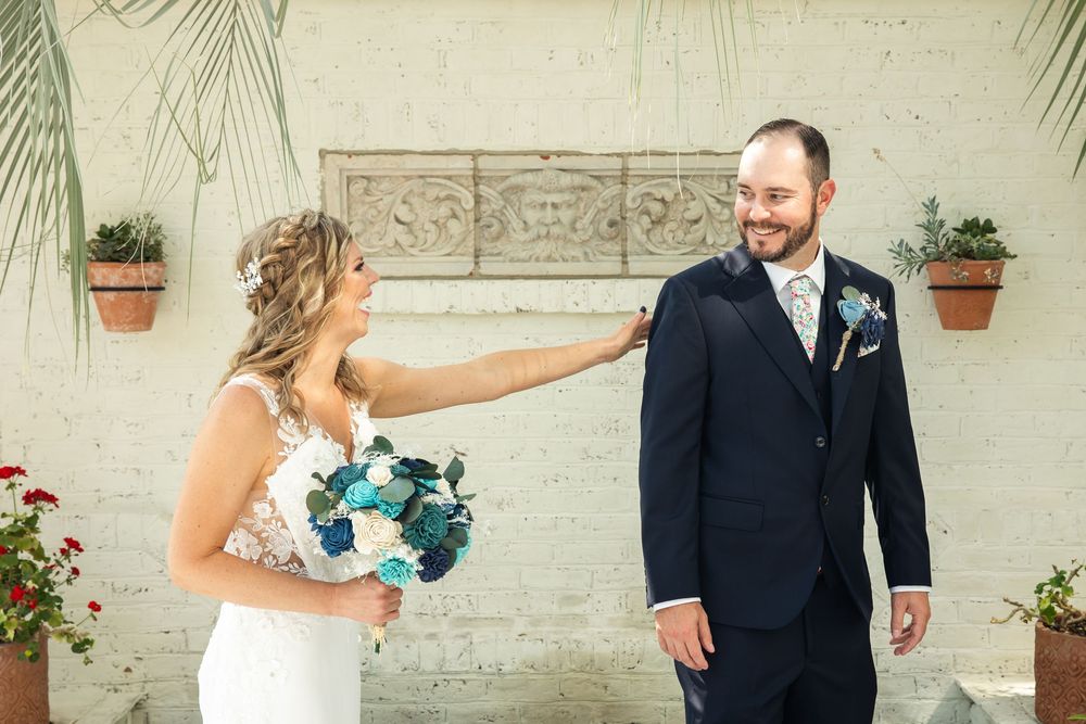 Bride in lace gown holds blue bouquet touching groom's shoulder in navy suit during romantic first look.