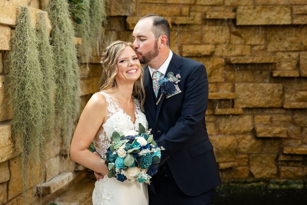Groom in navy suit kisses bride wearing lace gown holding blue bouquet beside stone wall with moss.