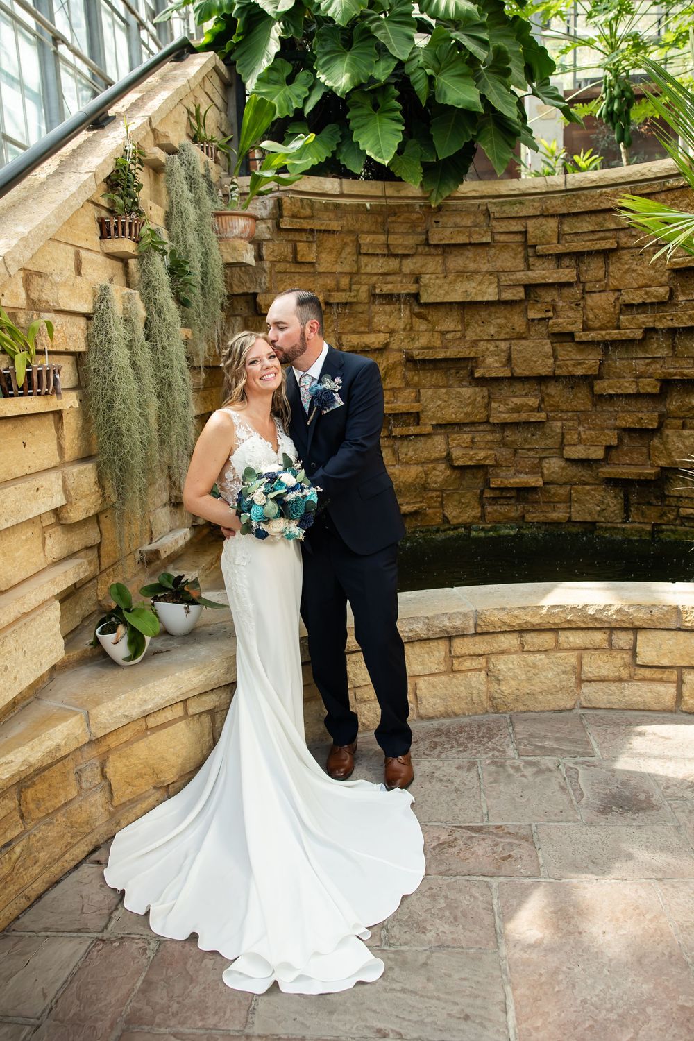 Bride and groom share a romantic kiss in a lush greenhouse setting, bride holding a bouquet in a white gown.