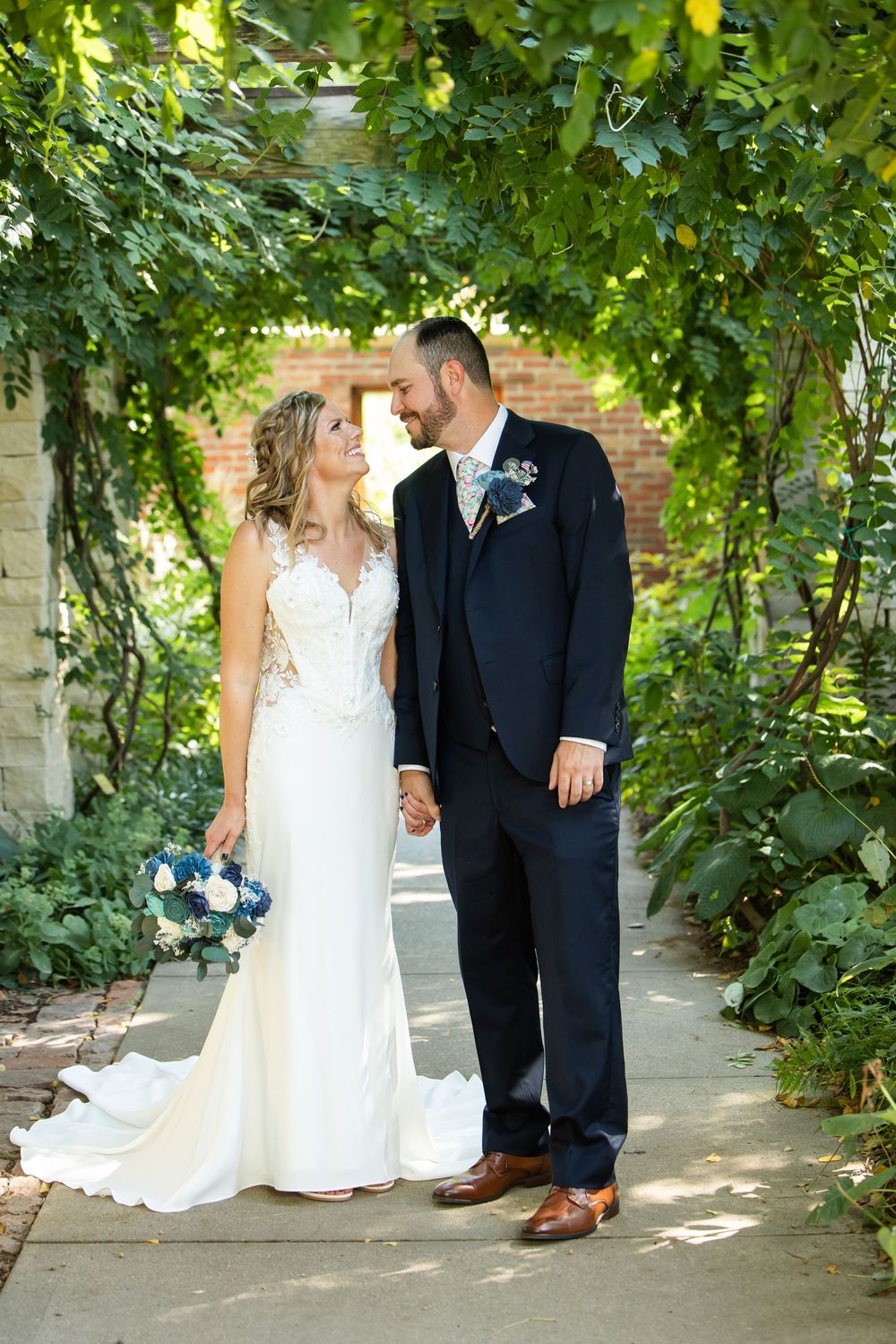 Bride and groom share a romantic moment under a lush green vine archway, bride holding a blue and white bouquet.