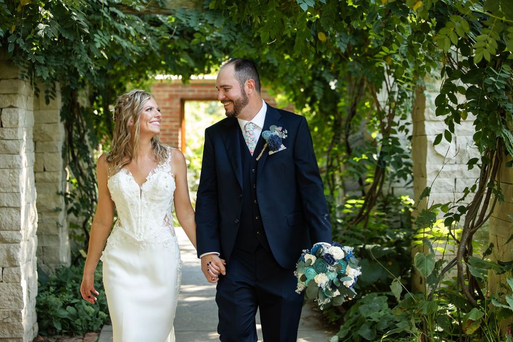 A bride and groom hold hands and smile at each other under a lush ivy-covered archway on their wedding day.