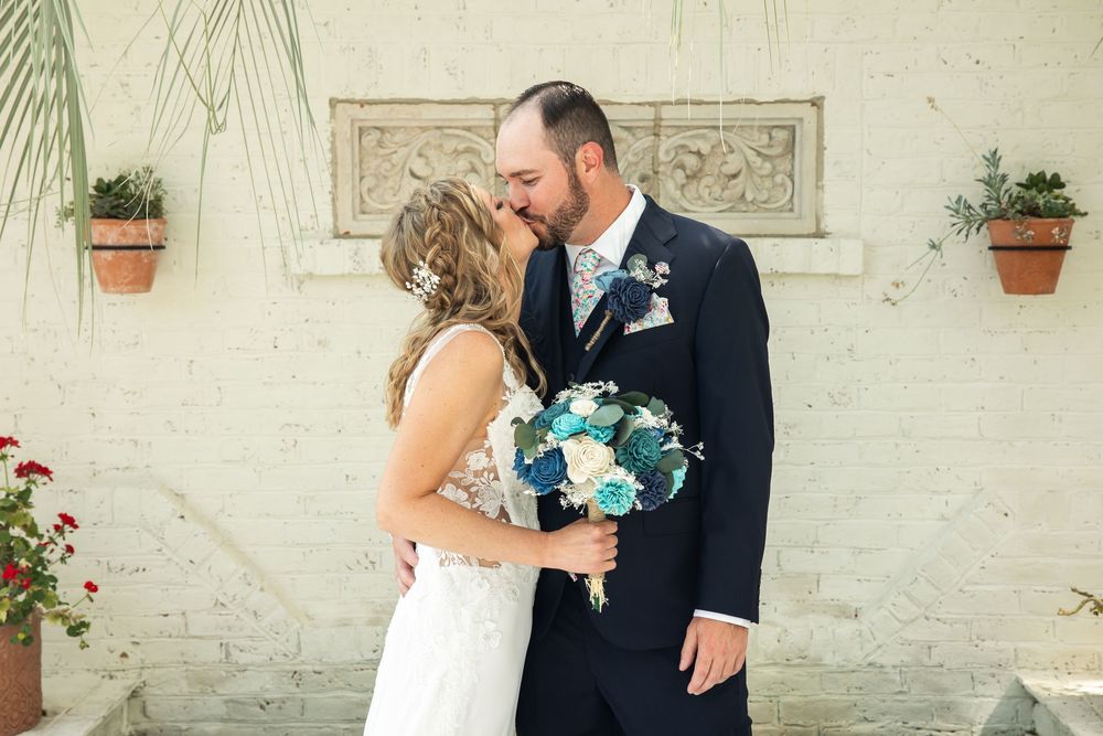 Bride and groom share a kiss outdoors, holding a blue and white bouquet, with a white brick wall and potted plants behind them.