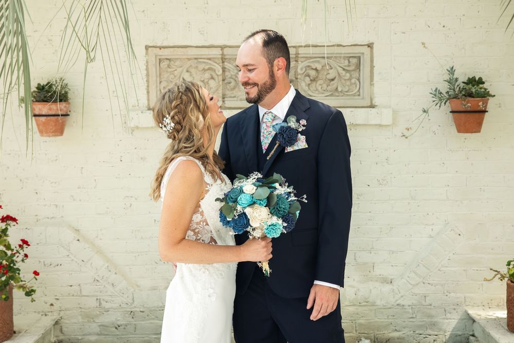 Bride and groom smiling at each other outdoors, holding a blue and white floral bouquet, groom in navy suit with floral tie.