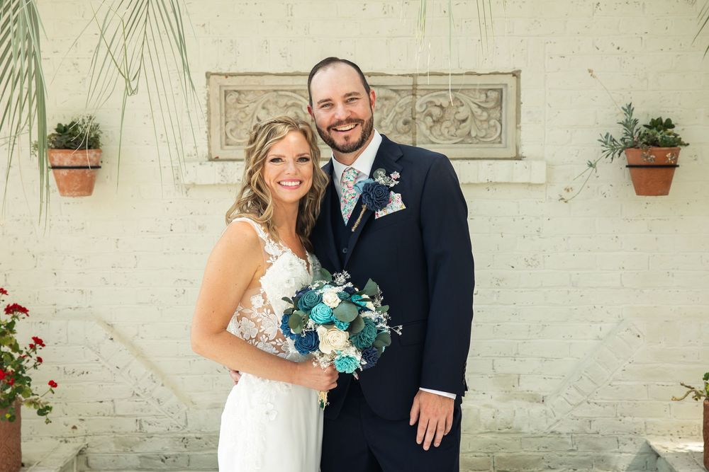Bride and groom smiling together outdoors, bride holding blue and white bouquet, groom in navy suit with floral tie.