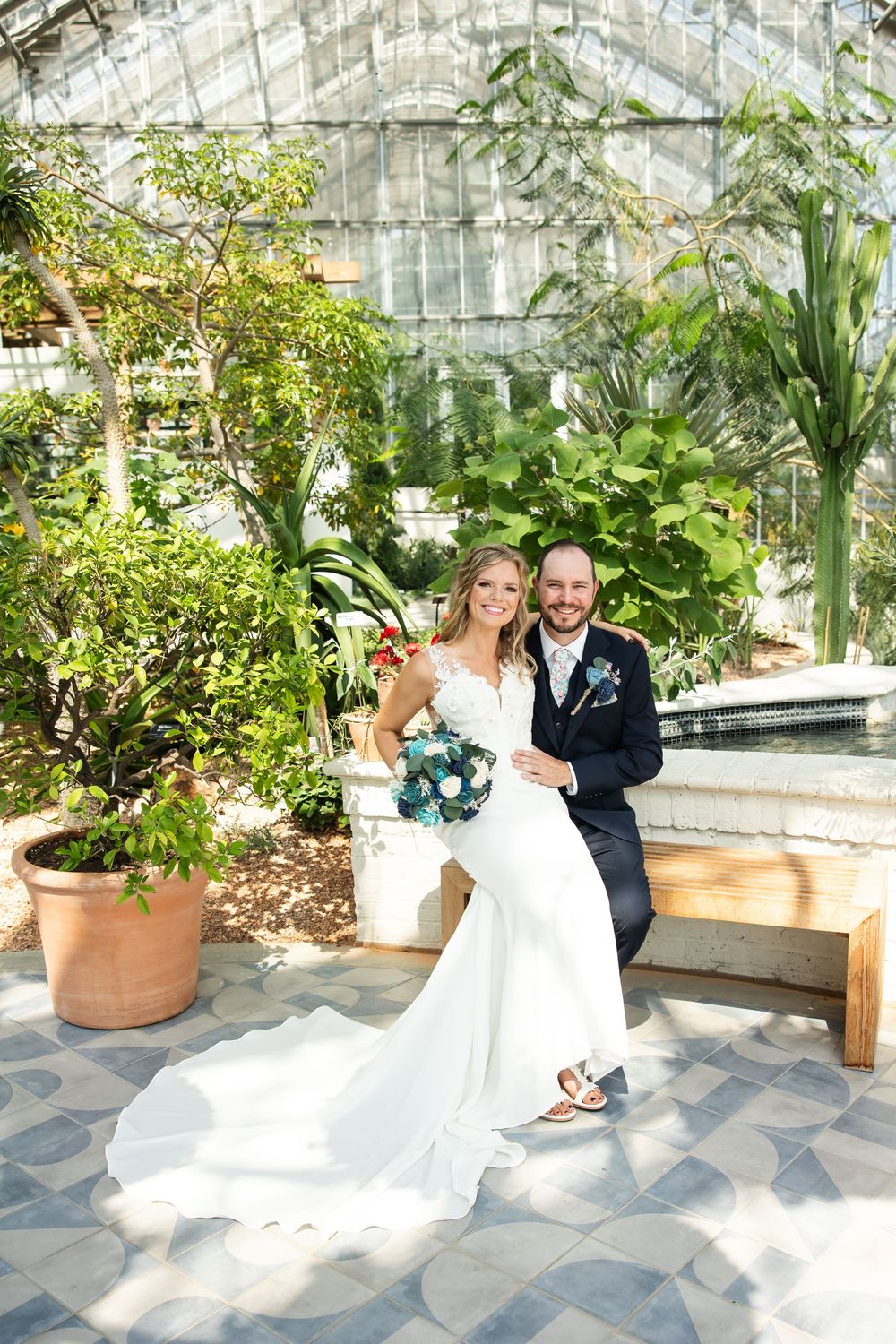Bride and groom sitting together on a bench in a lush greenhouse, bride holding a blue bouquet in a white gown.