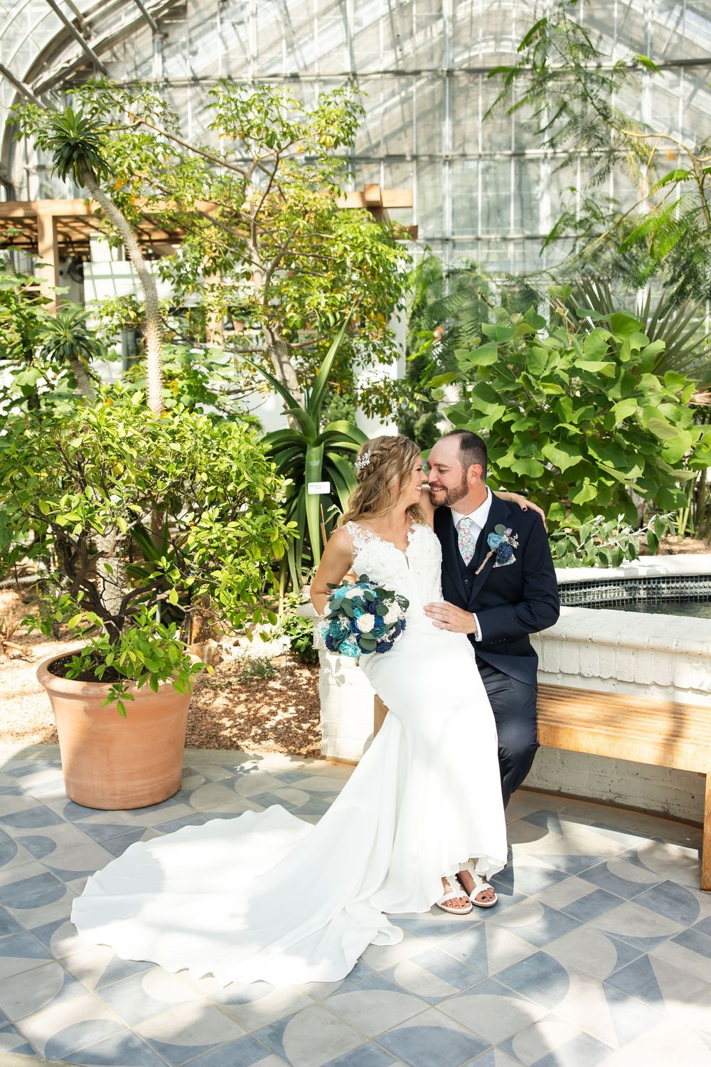 Bride and groom share a romantic moment in a sunlit greenhouse, surrounded by lush tropical plants.