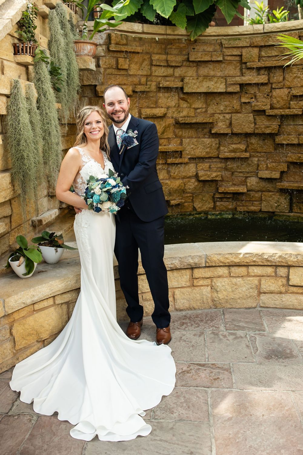 Bride in white gown holding teal bouquet poses with groom in navy suit before stone wall fountain.