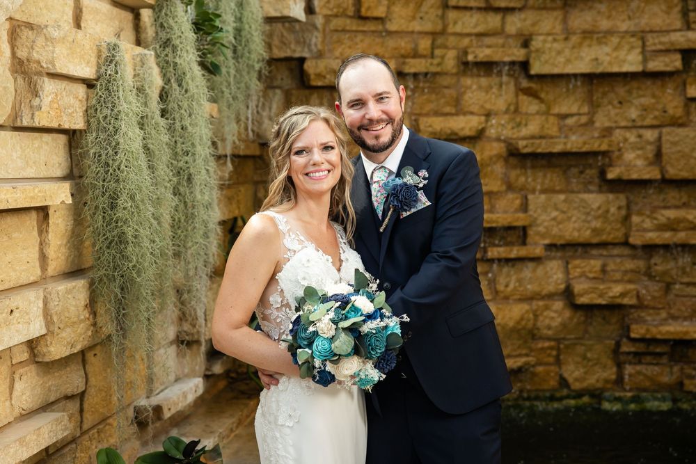 Smiling bride holding teal bouquet stands with groom in navy suit before stone wall with greenery.