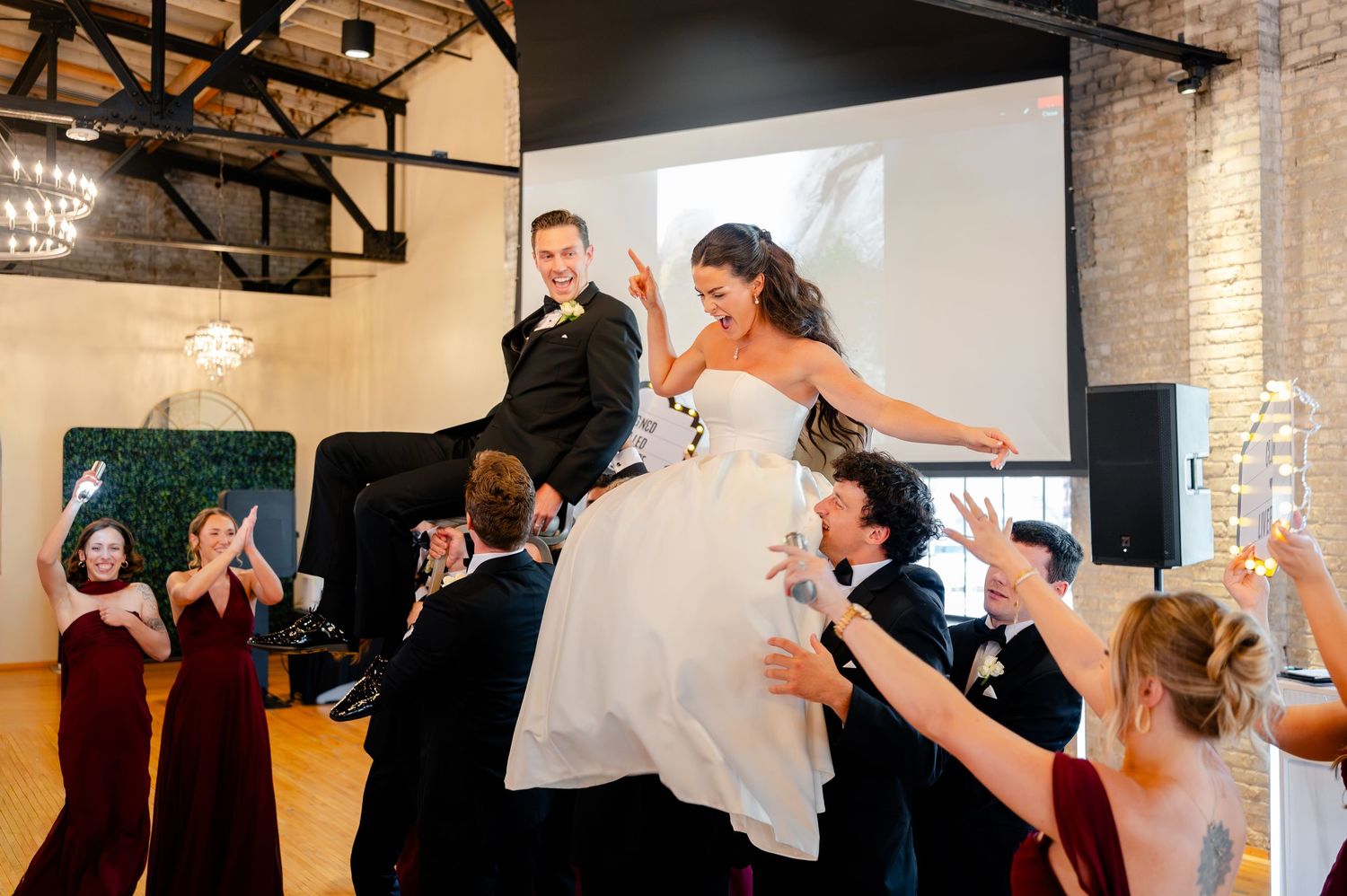 Bride and groom lifted by wedding party during joyful reception celebration in rustic industrial venue.