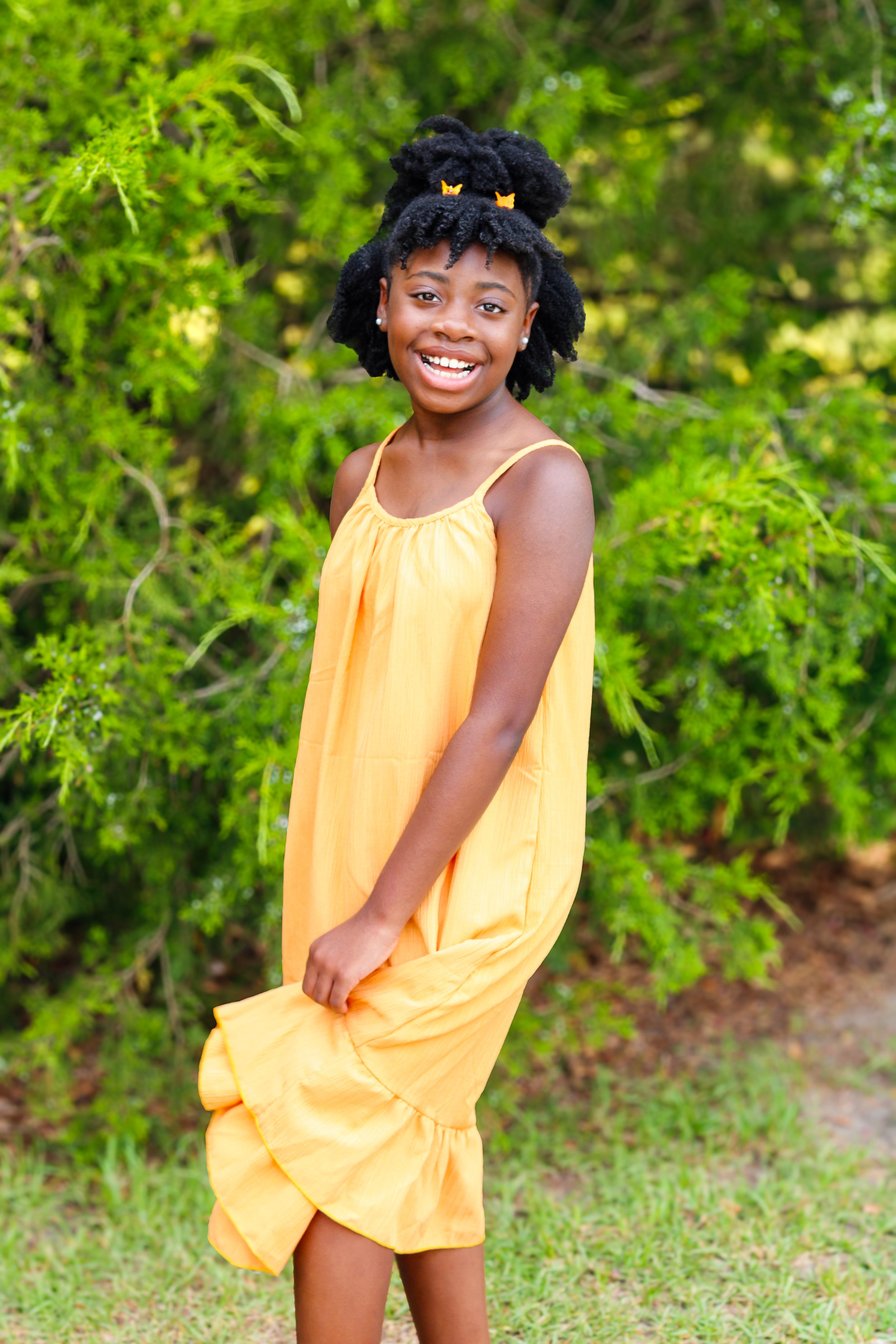 Smiling young Black girl in yellow sundress with butterfly hair clips poses outdoors among lush green trees.