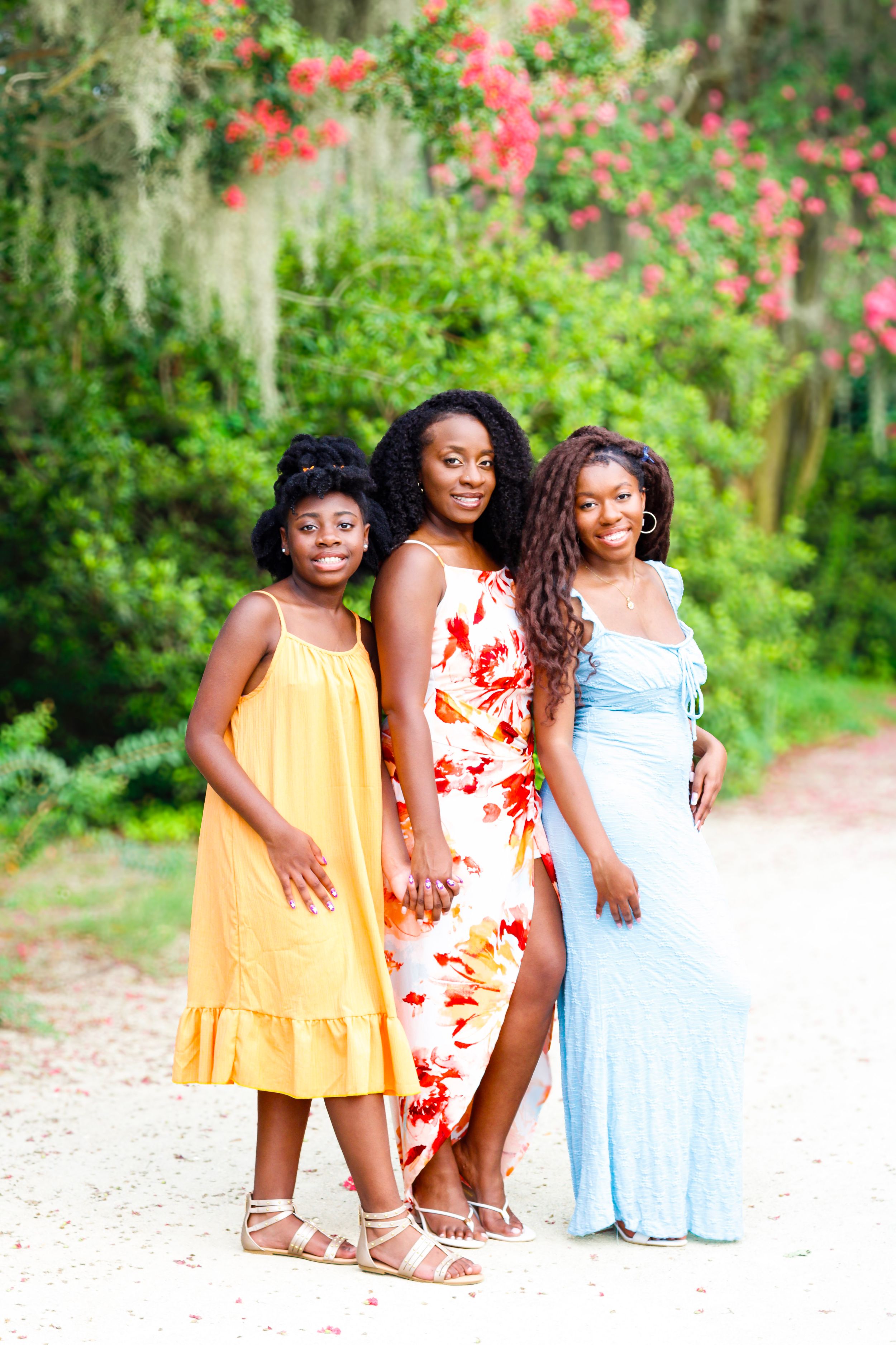 Three young Black women in colorful dresses pose together on a sandy path surrounded by lush green trees and pink flowers.
